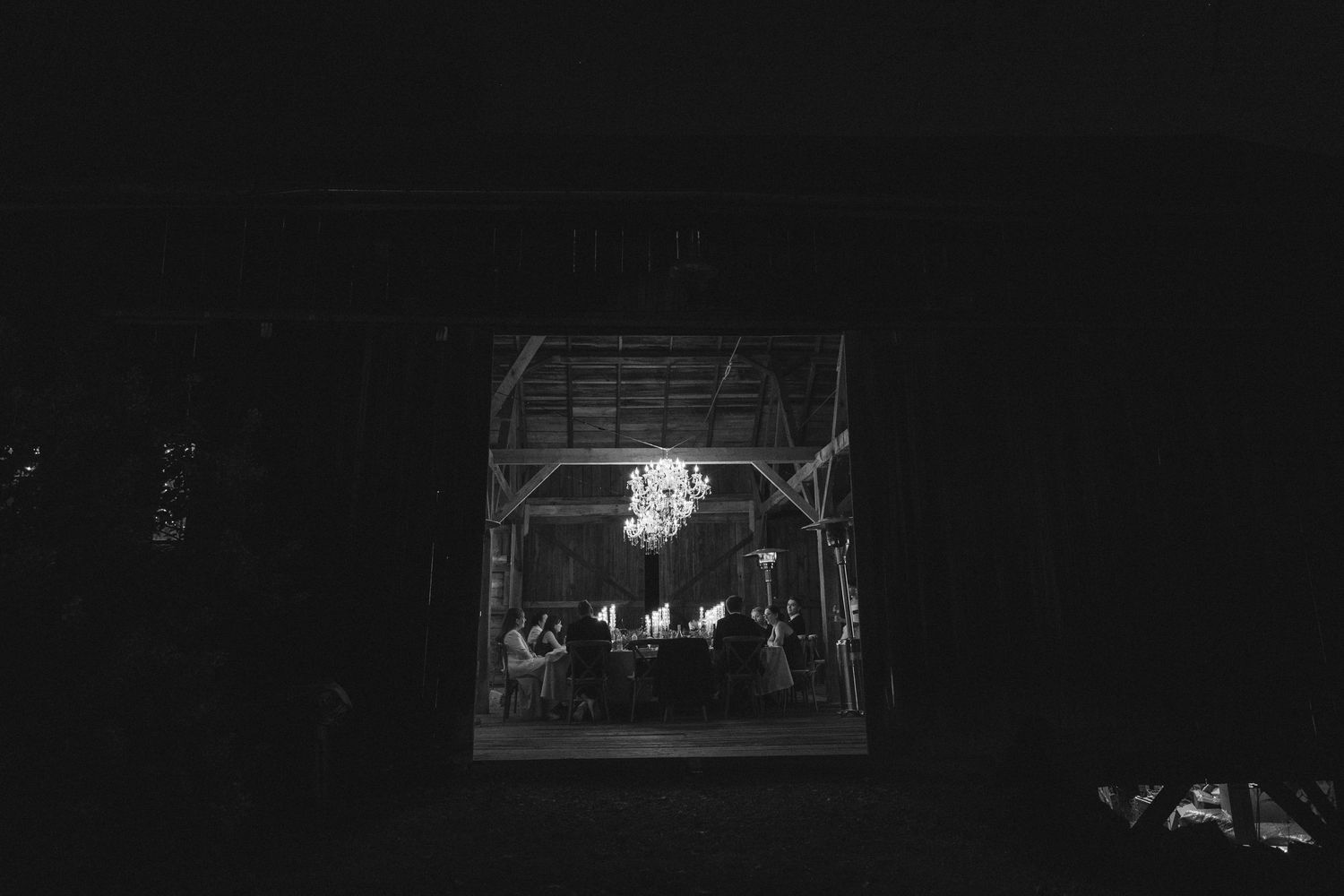 Black and white barn wedding reception with chandelier lighting and guests seated at tables in rustic venue.