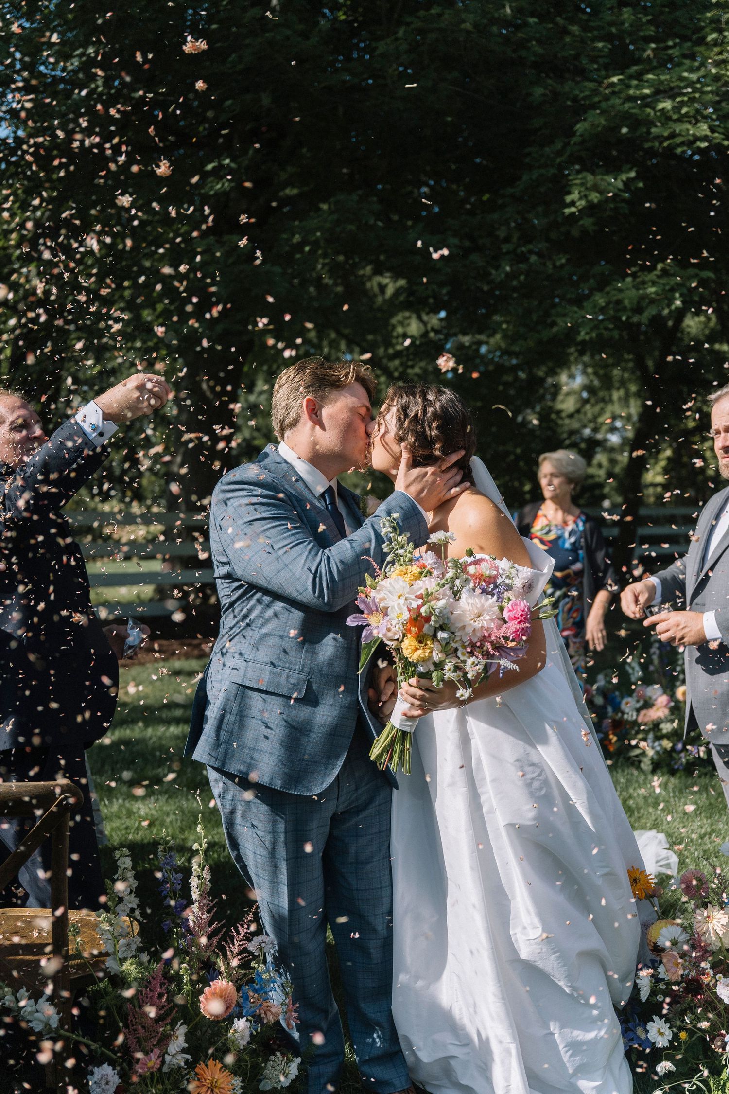 Wedding couple celebrates with confetti toss in outdoor garden setting while embracing during summer celebration.