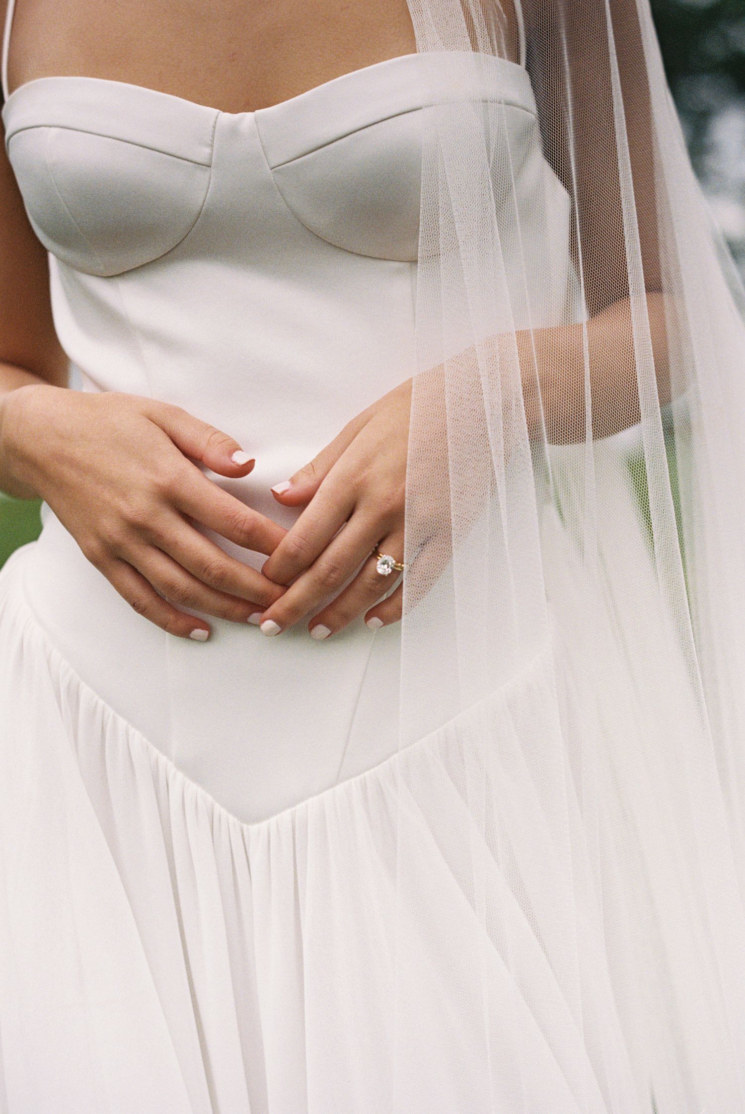 Bride in white strapless wedding gown with tulle veil, hands clasped at waist, diamond engagement ring visible.