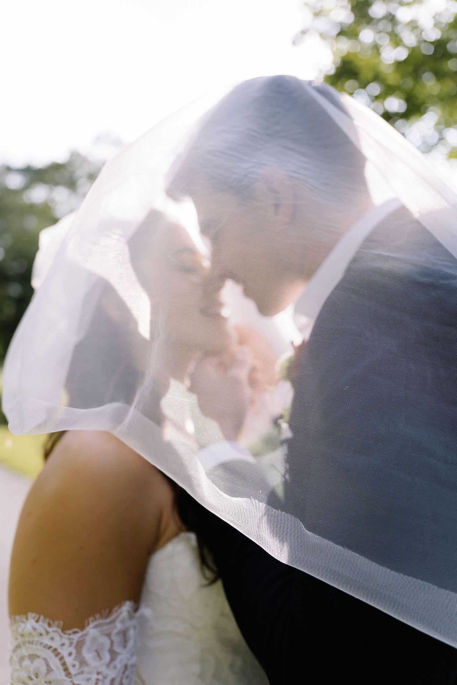 A romantic wedding moment captured through a flowing white veil as newlyweds share an intimate kiss in sunlit garden setting.