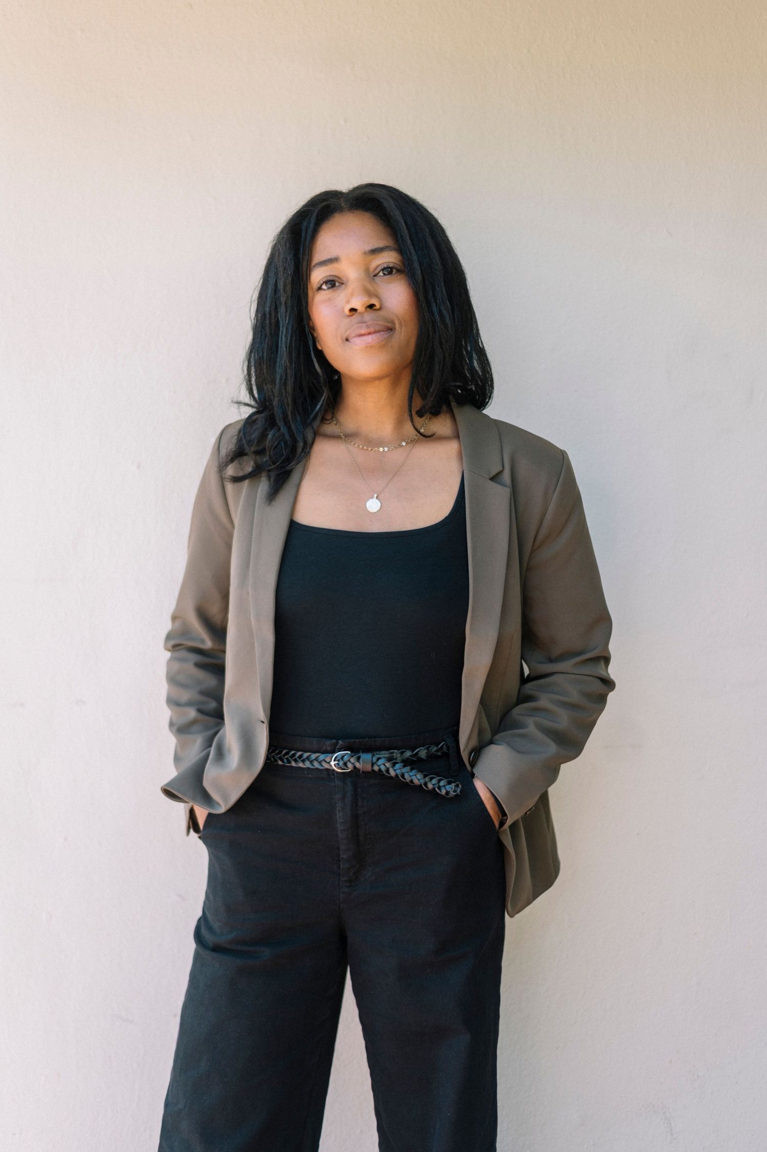 A young Black woman in a olive blazer, black top, and pants stands confidently with hands in pockets against a neutral wall.