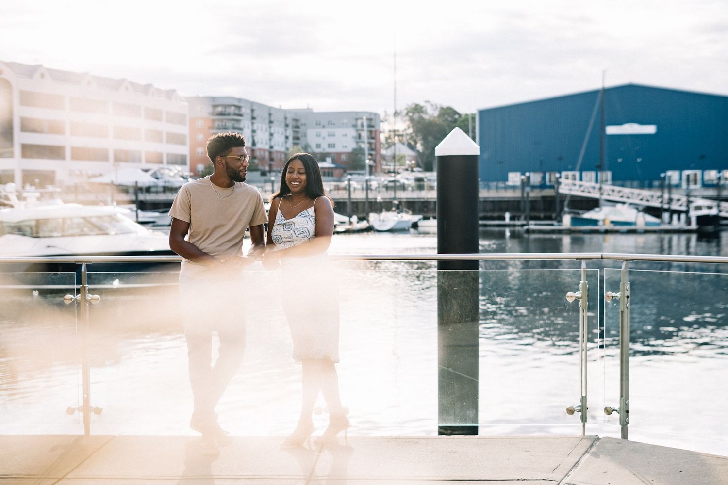 A couple stands on a wooden dock overlooking a marina filled with boats on a bright sunny day.