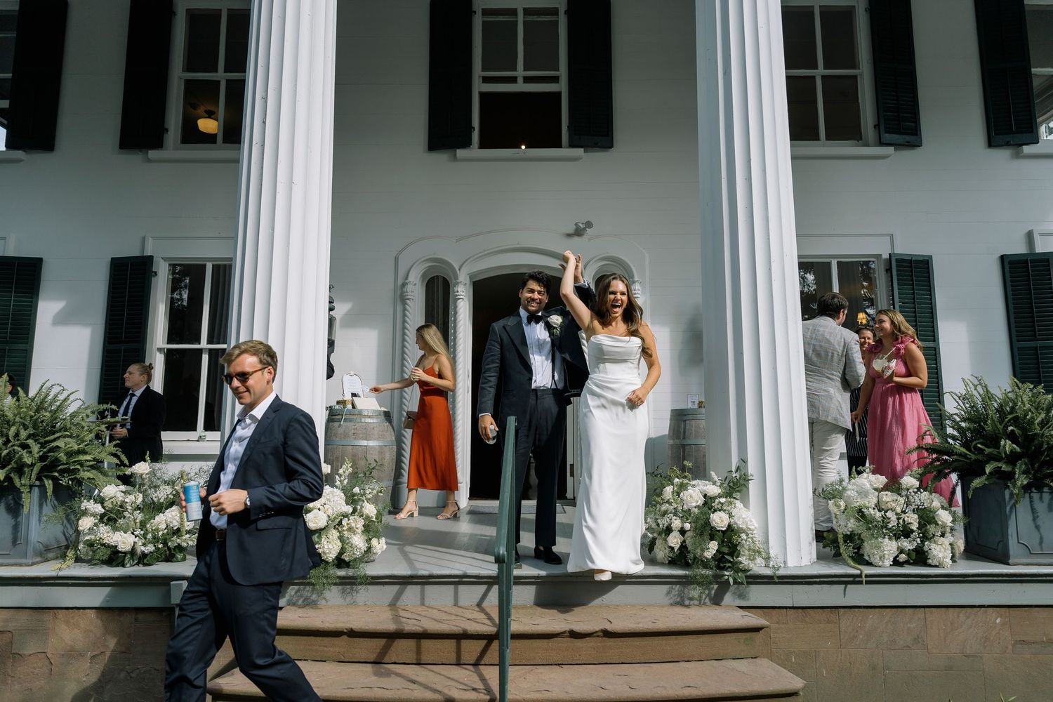 A festive wedding couple celebrates with their bridal party on the porch of a grand white-columned building.