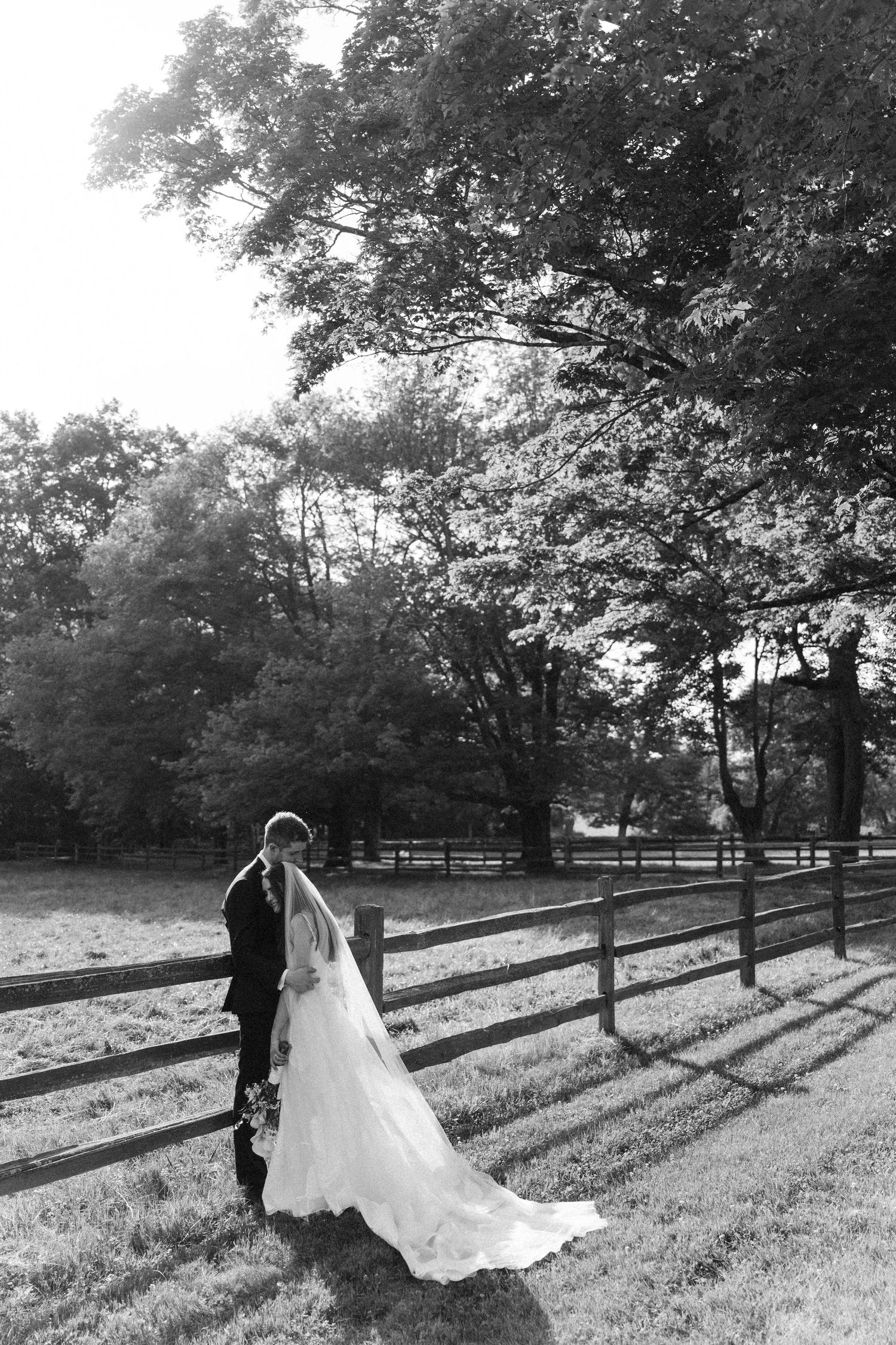 Black and white photo of a bride and groom kissing near a wooden fence surrounded by lush trees on their wedding day.