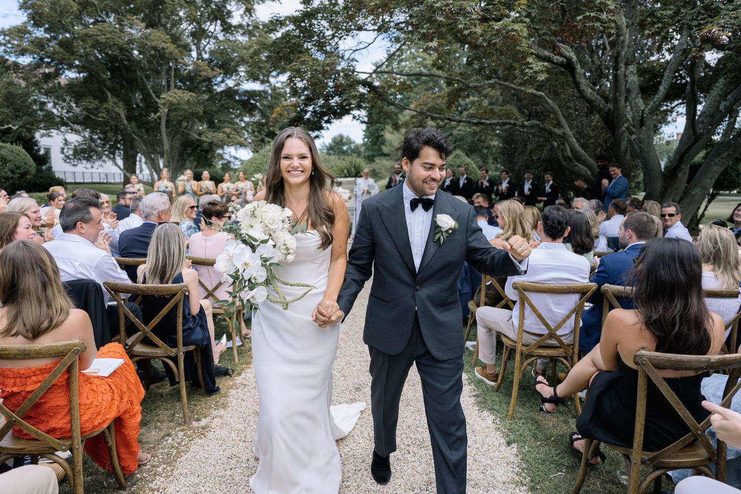 Wedding guests mingle and celebrate at an outdoor reception with rustic wooden chairs and natural decor.