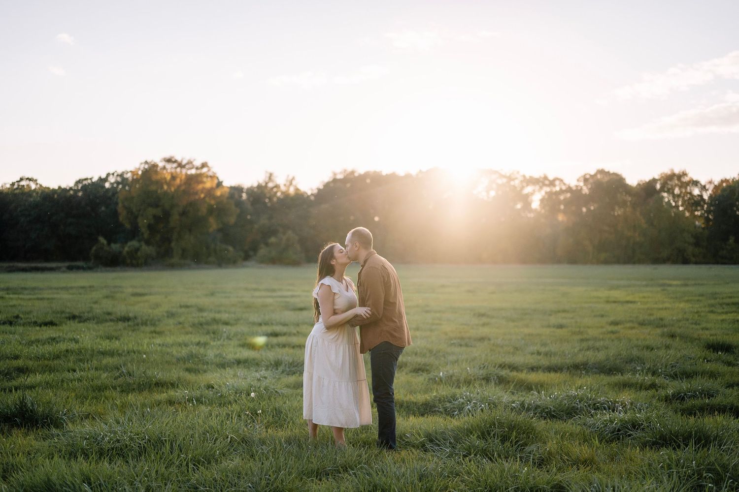 A couple embraces in a sunlit meadow during golden hour with trees silhouetted in the background.