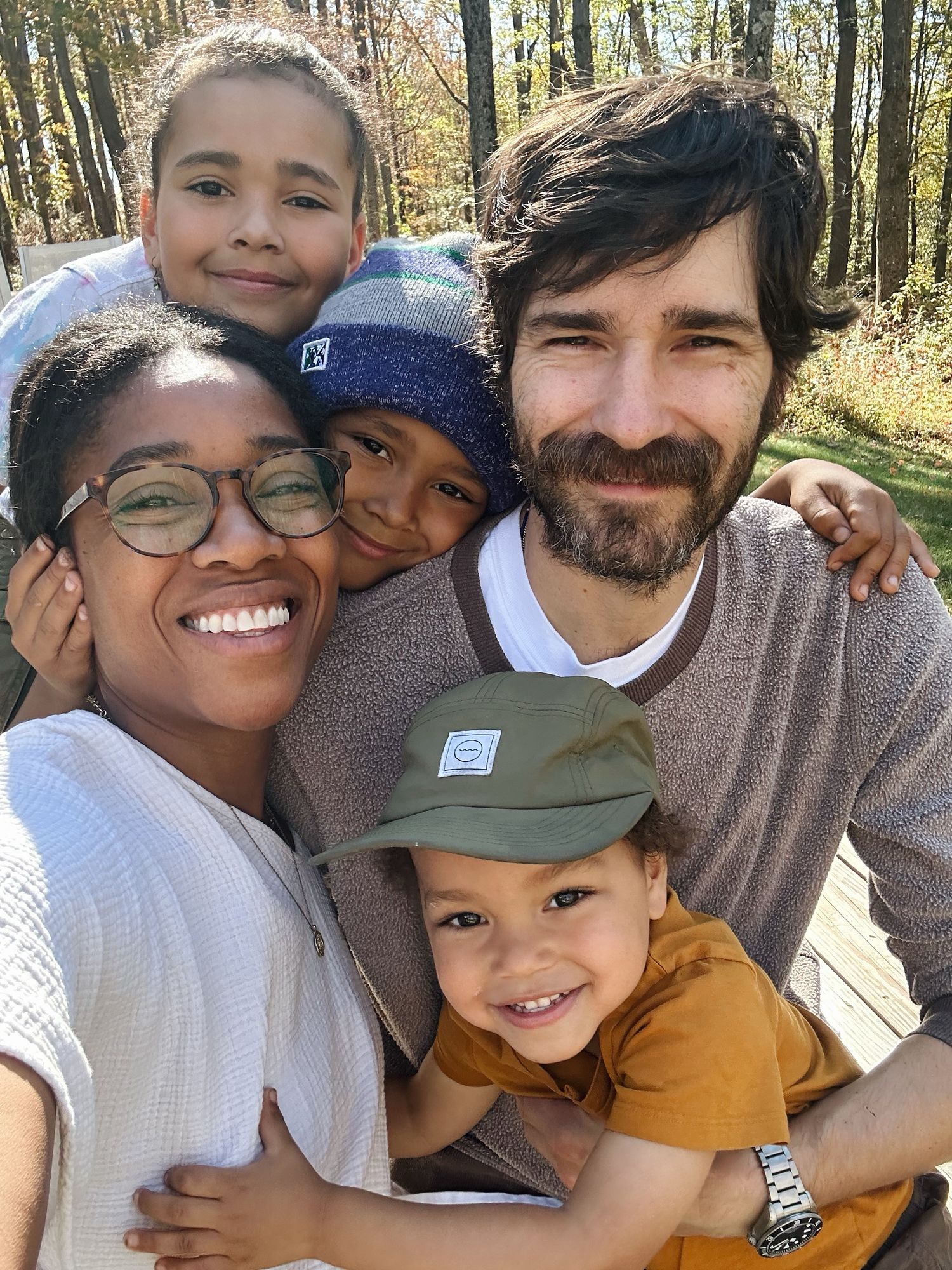 Happy family of five smiling together outdoors in autumn forest, parents with three young children taking a selfie.