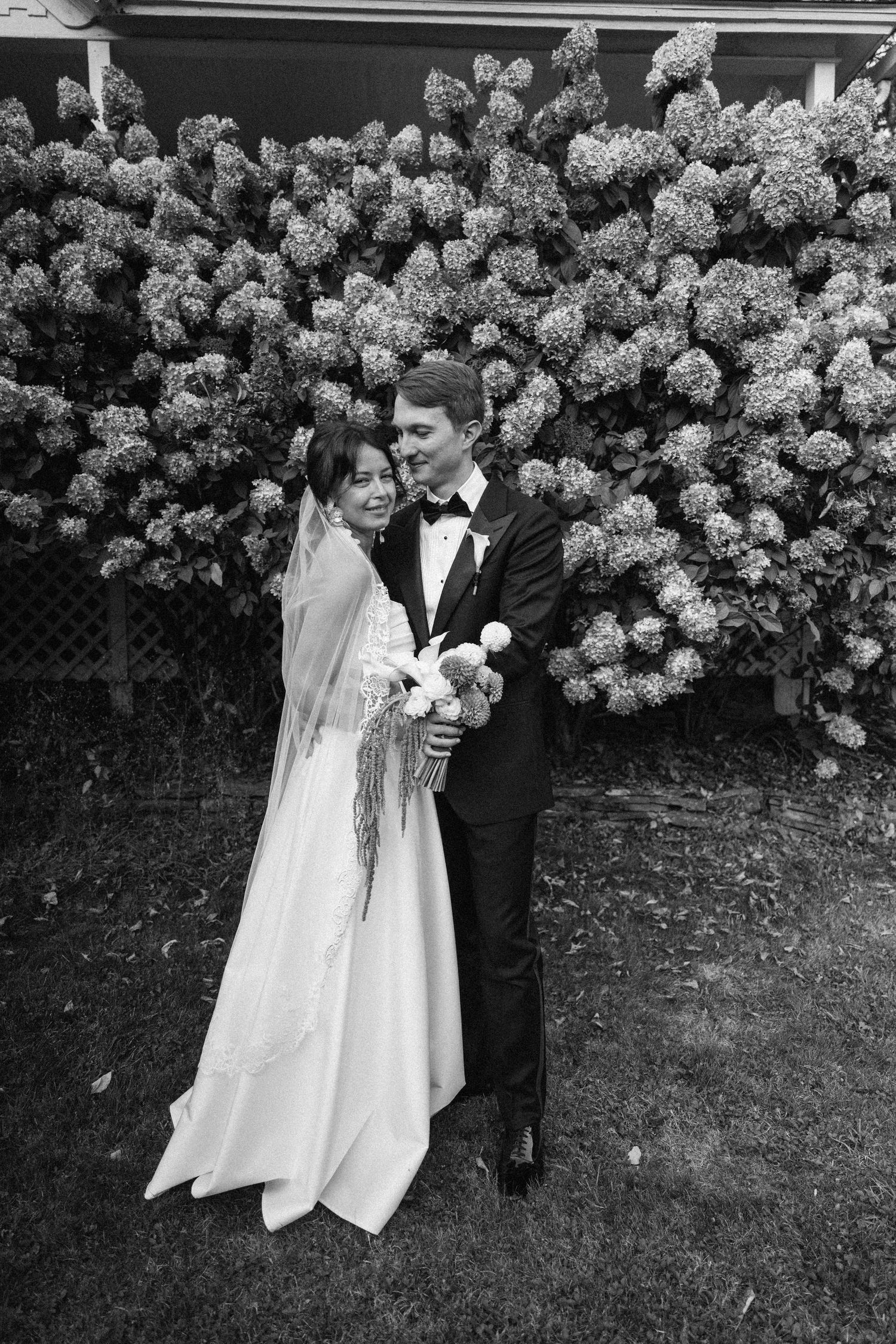 Black and white wedding photos showing couple in formal attire posing together against flowering hydrangea bush backdrop.