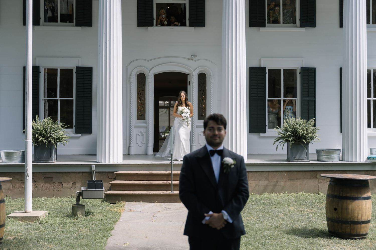 A wedding party stands in front of a white columned mansion with barrels and greenery decorating the entrance.