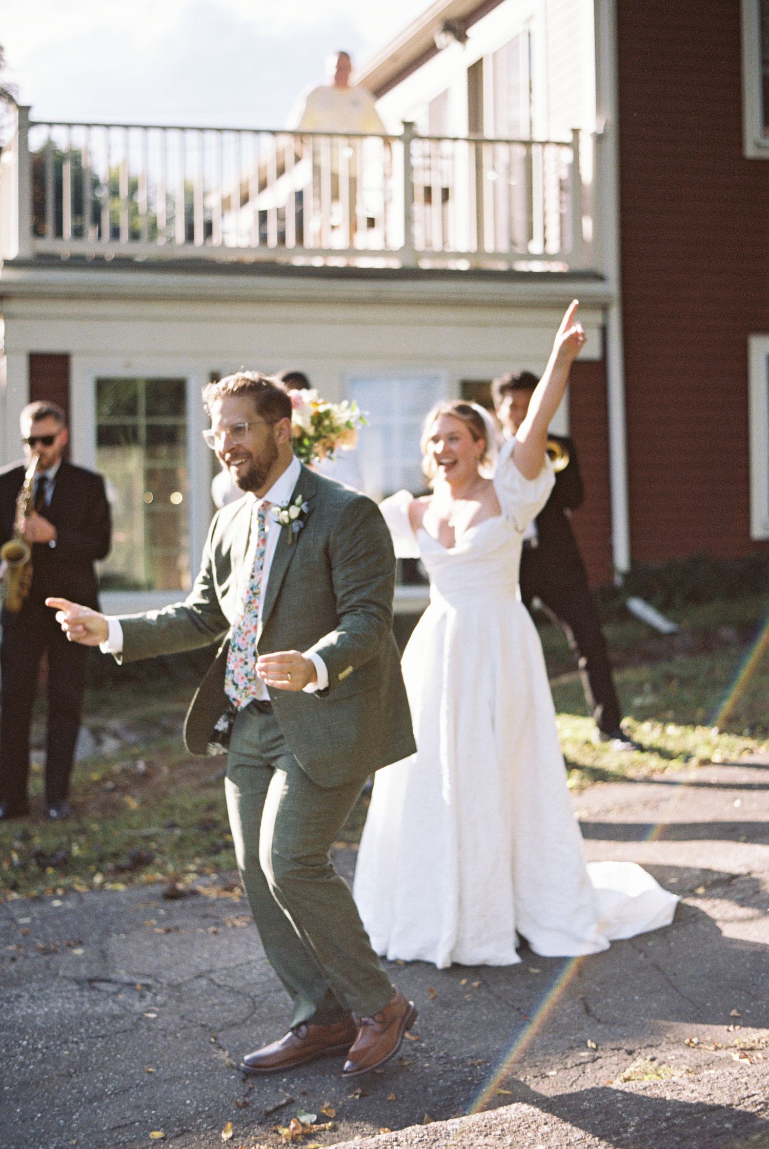 Joyful bride in a white gown and groom in a green suit dance outdoors at their wedding as a saxophonist plays behind them.