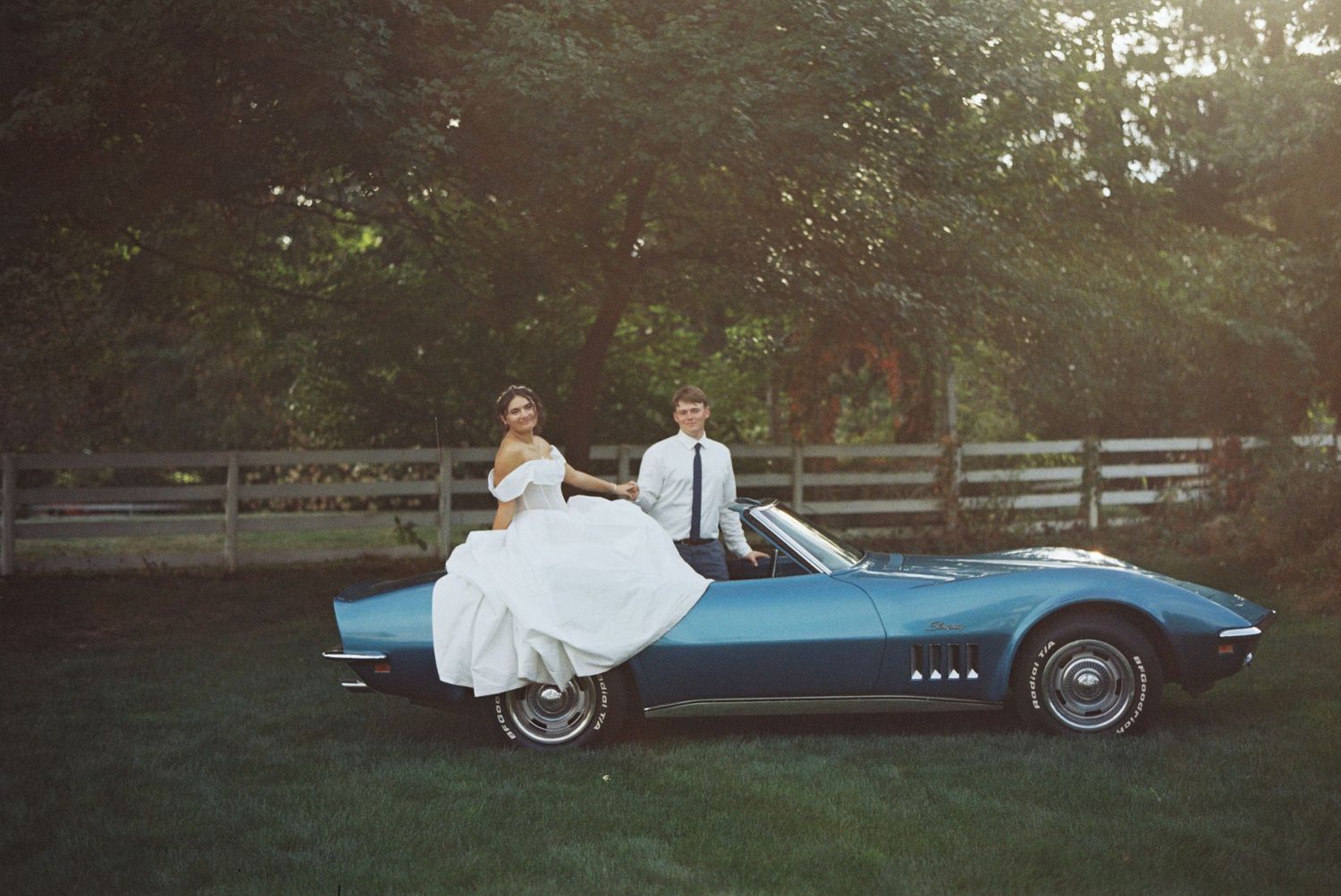 Bride and groom posing with a classic blue Corvette convertible on a green lawn at sunset.