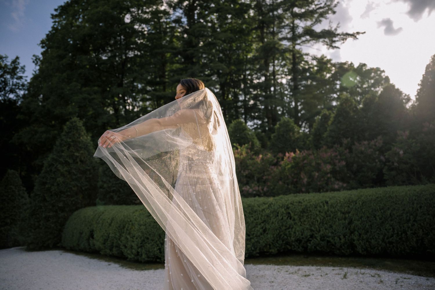 A flowing white veil catches the breeze in a garden setting during sunset, creating an ethereal bridal moment.