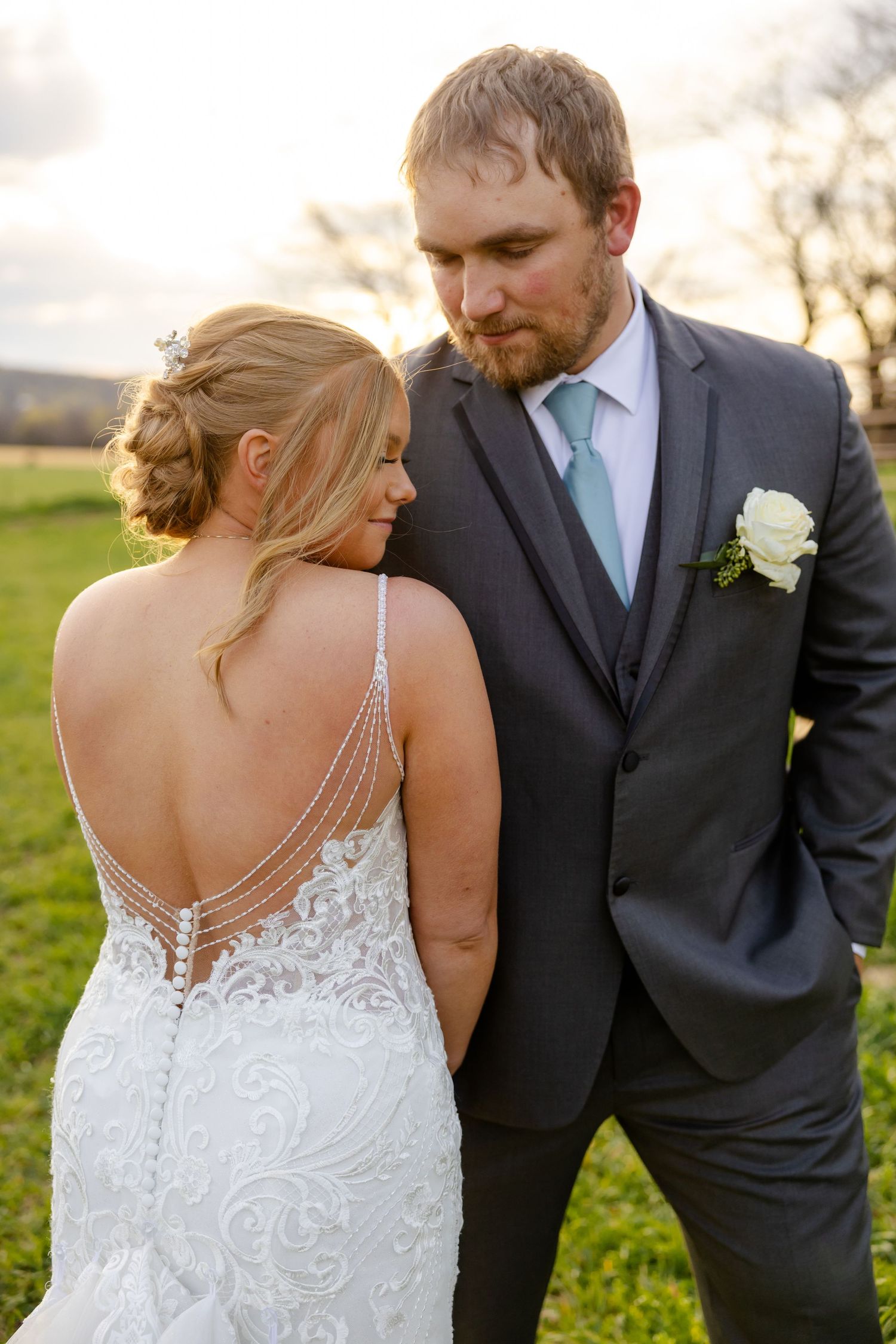 A bride in an intricate lace back dress stands close to a groom in a dark suit with a mint tie during an outdoor wedding.