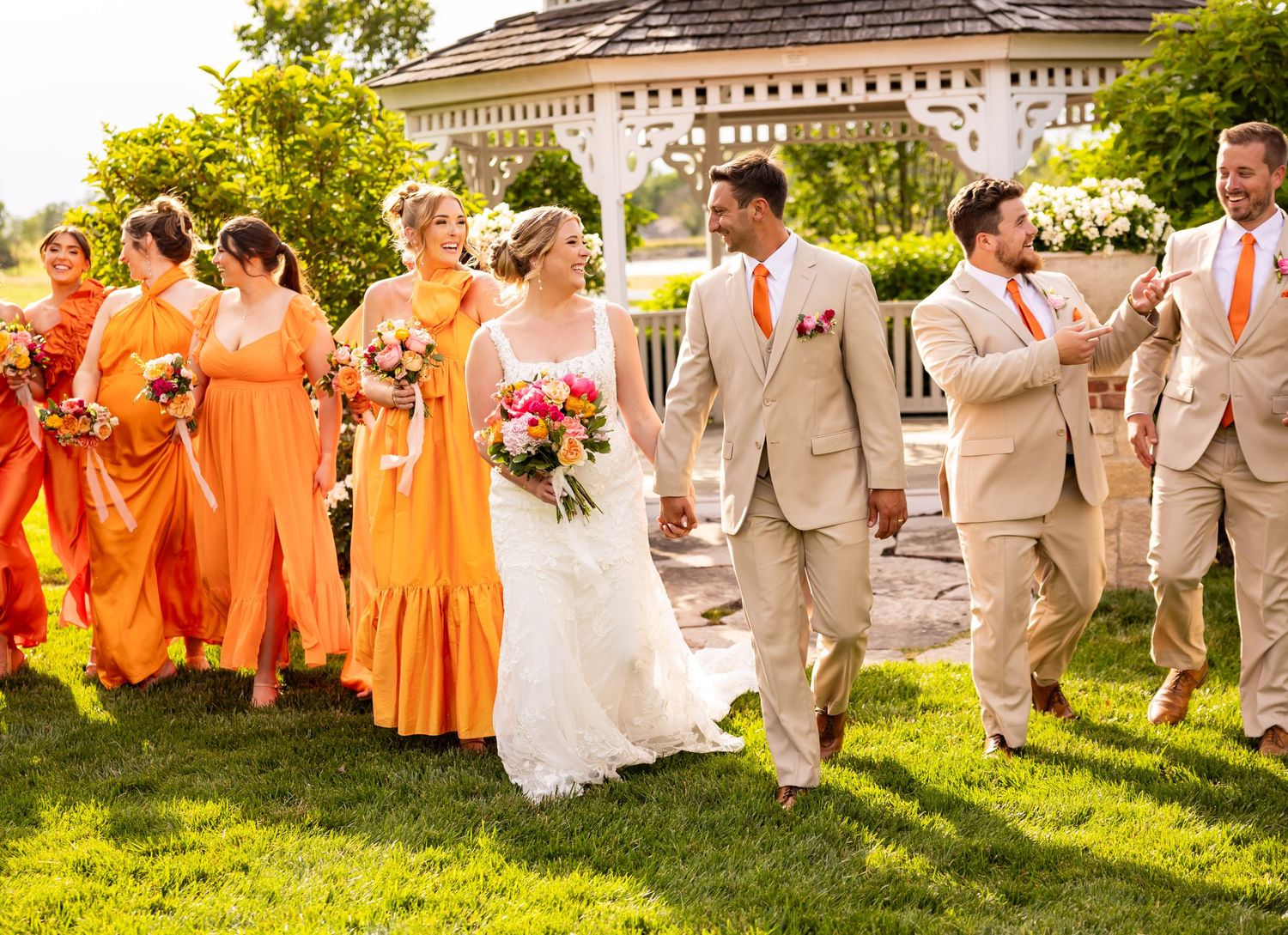 A wedding party wearing orange dresses and beige suits walks together on grass near an ornate building on a sunny day.