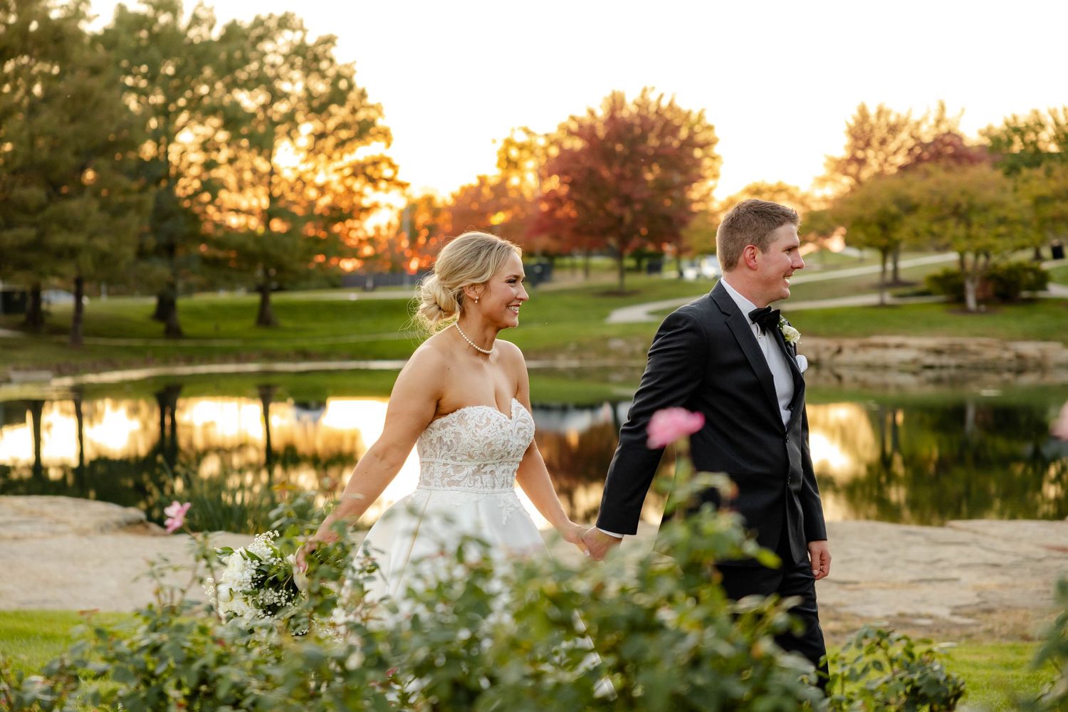 A romantic sunset wedding photo by a serene pond with golden light filtering through autumn trees.