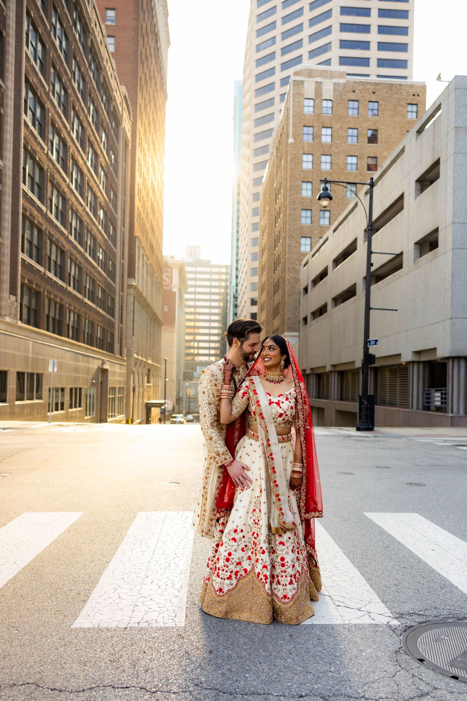 A couple in traditional wedding attire embrace on a city crosswalk at sunset with tall buildings in the background.