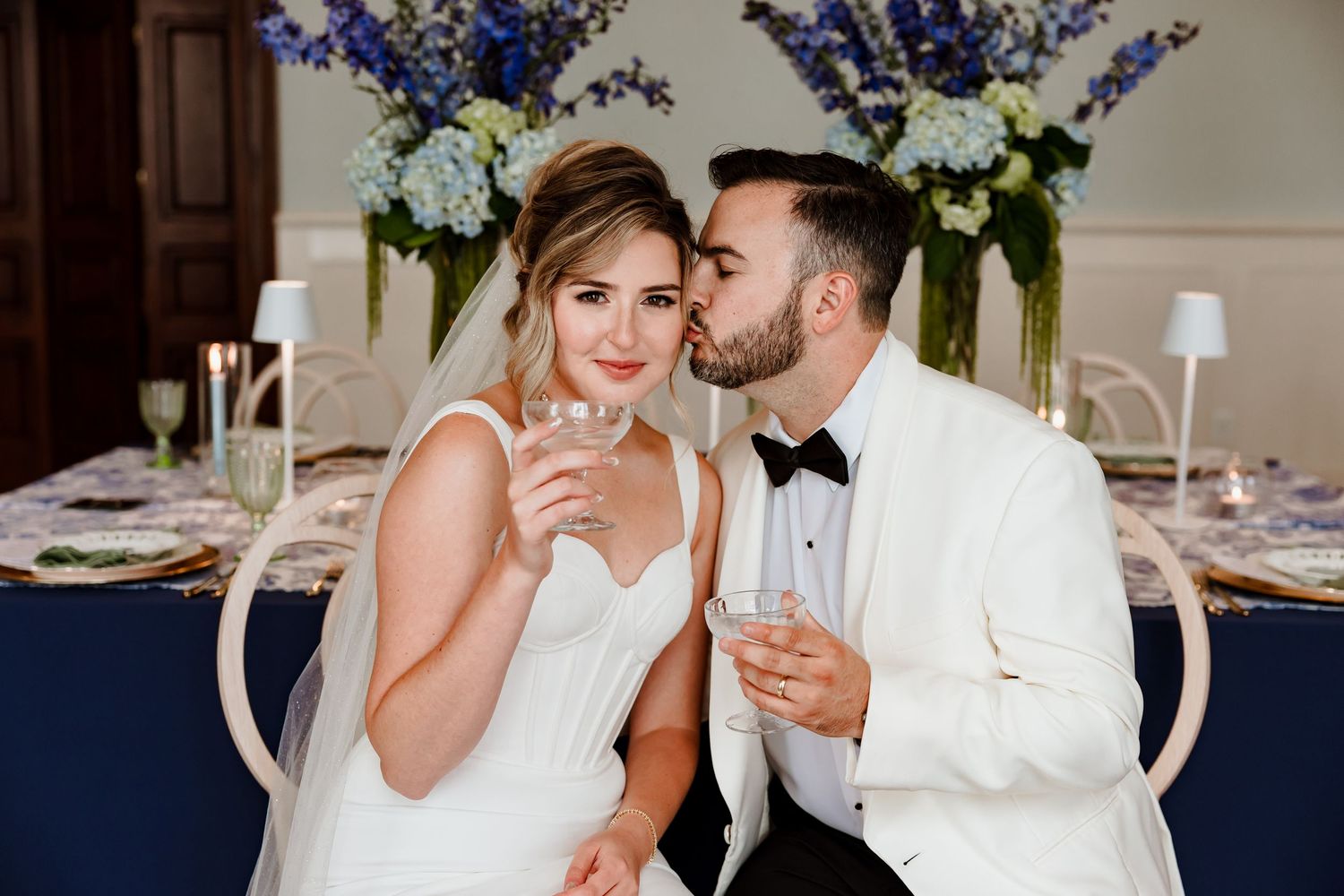 Couple in elegant wedding attire share champagne while sitting at a reception table with blue and white decor.