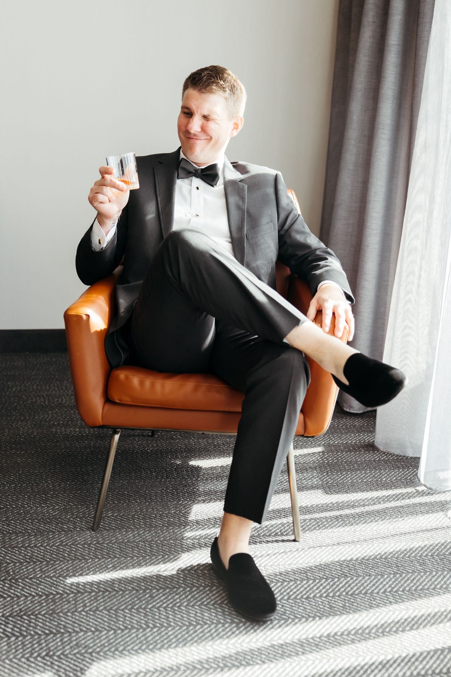 Elegant groom in black tuxedo with bow tie relaxes in leather chair while holding drink before wedding celebration.