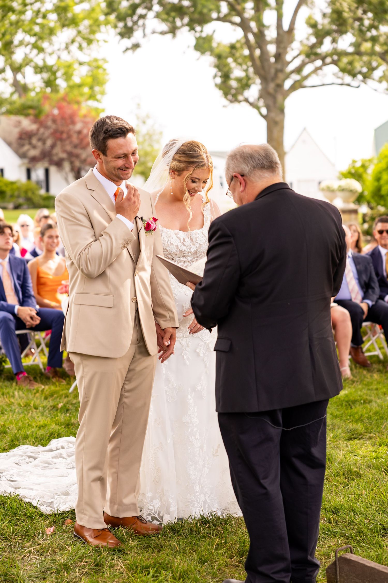An outdoor wedding ceremony takes place on a sunny day with guests seated on the lawn while the couple exchanges vows.