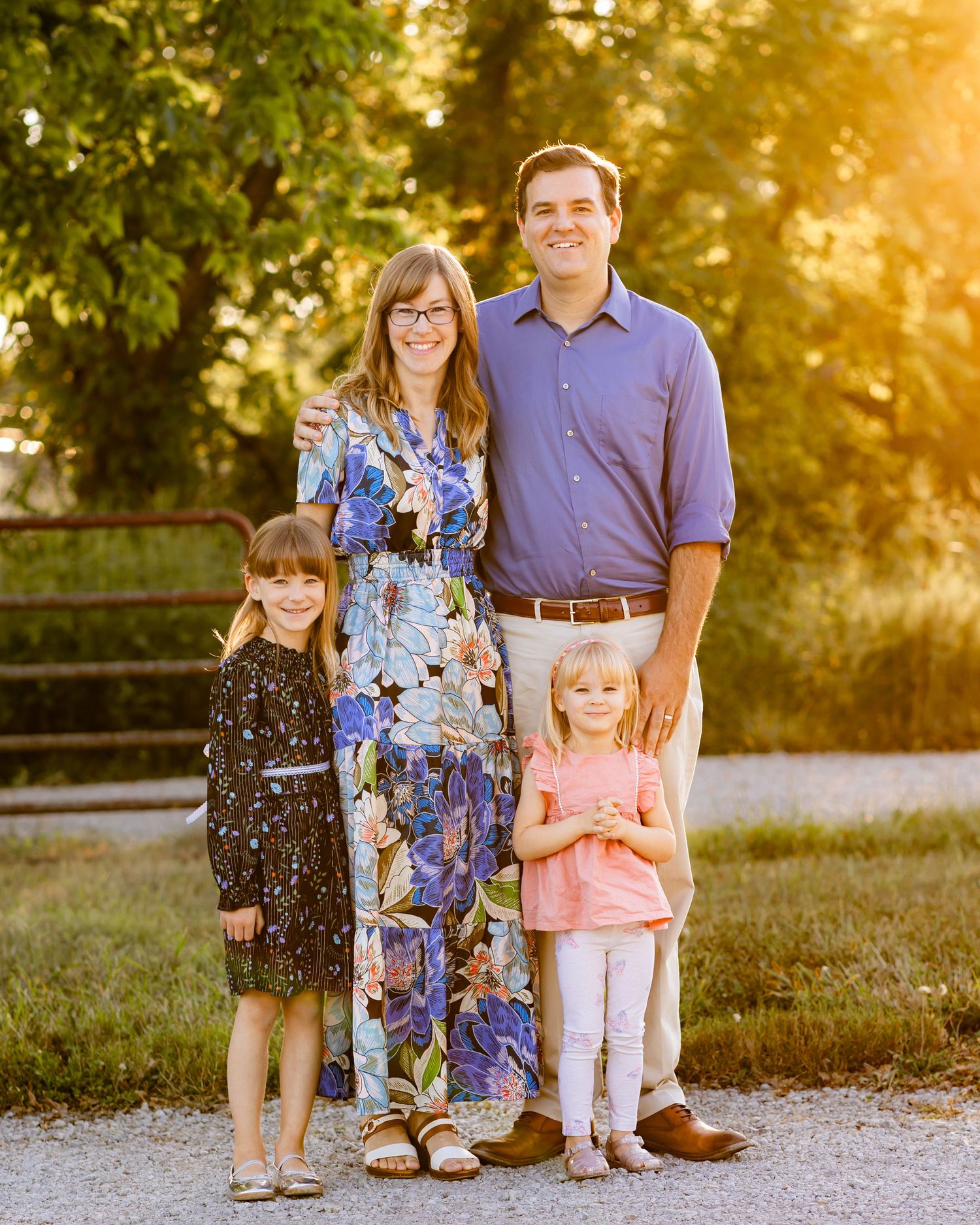 Family of four standing together on a gravel path with golden sunlight and trees in the background.