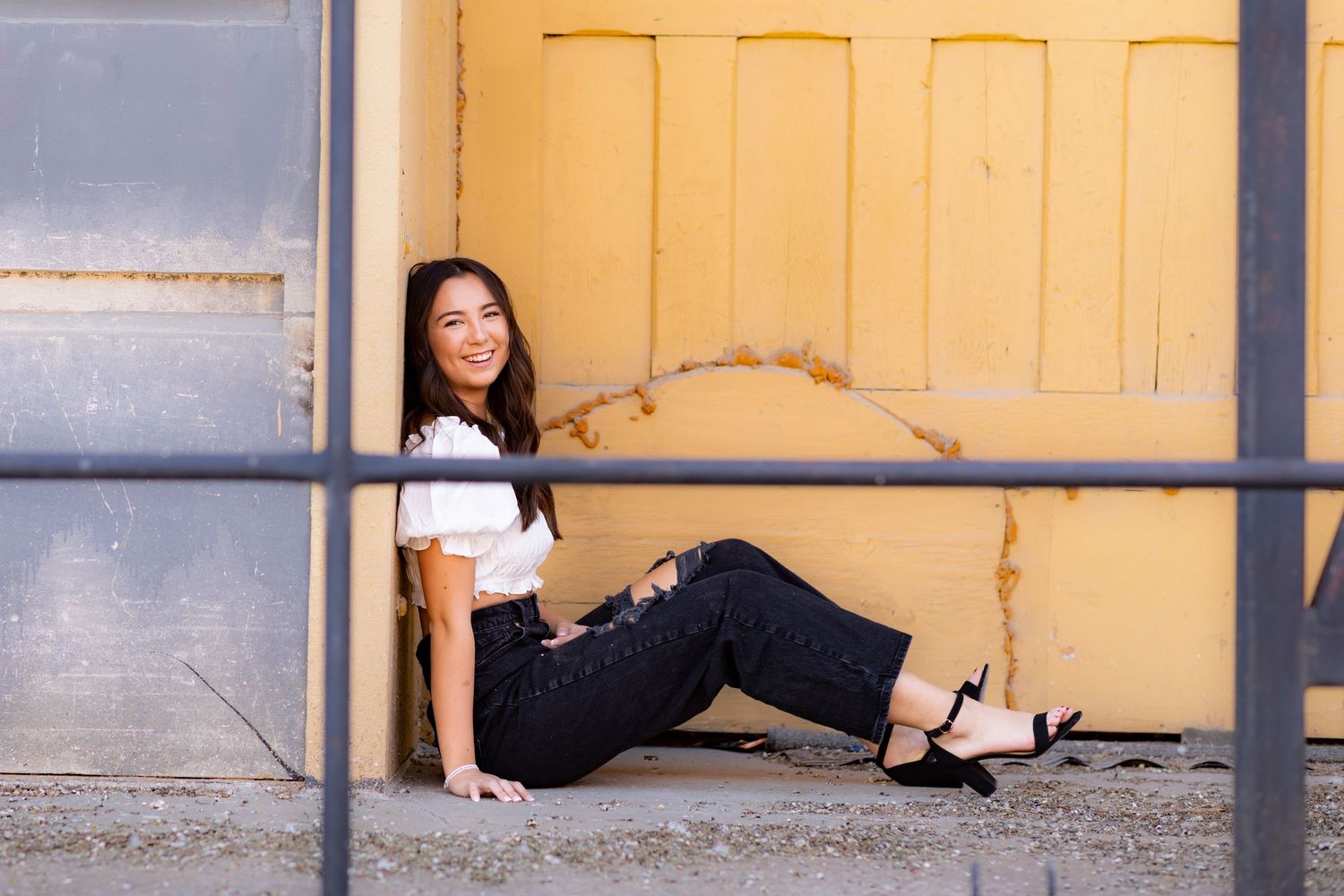 A casually dressed woman sits on the floor against a yellow door in an urban setting.