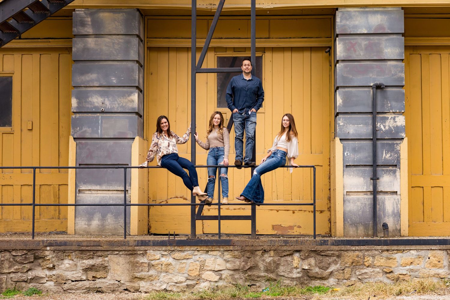 Group of people posing on a rustic yellow building porch with stone foundation and gray pillars.