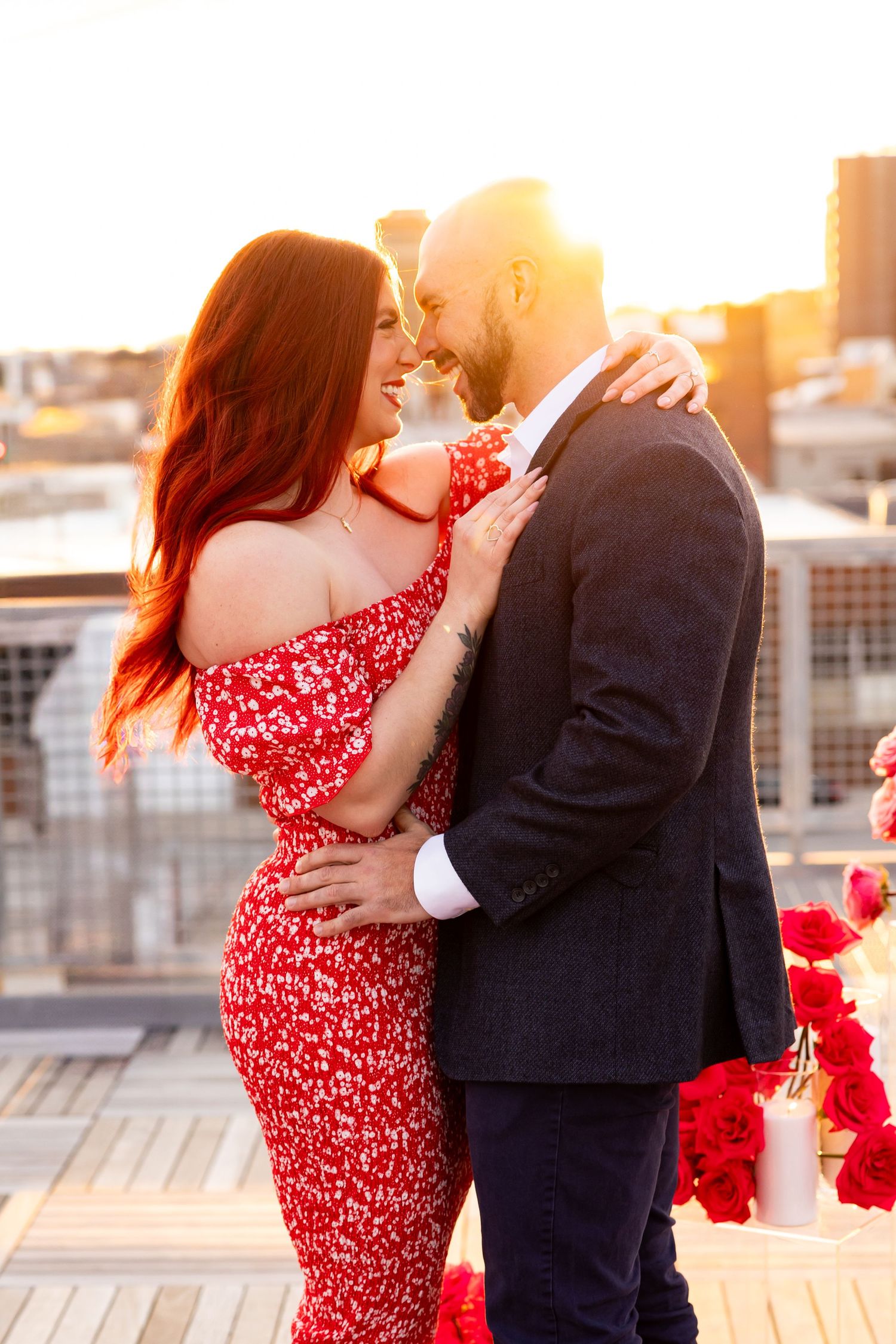 Couple in romantic embrace with woman wearing red floral dress during golden sunset on rooftop terrace.