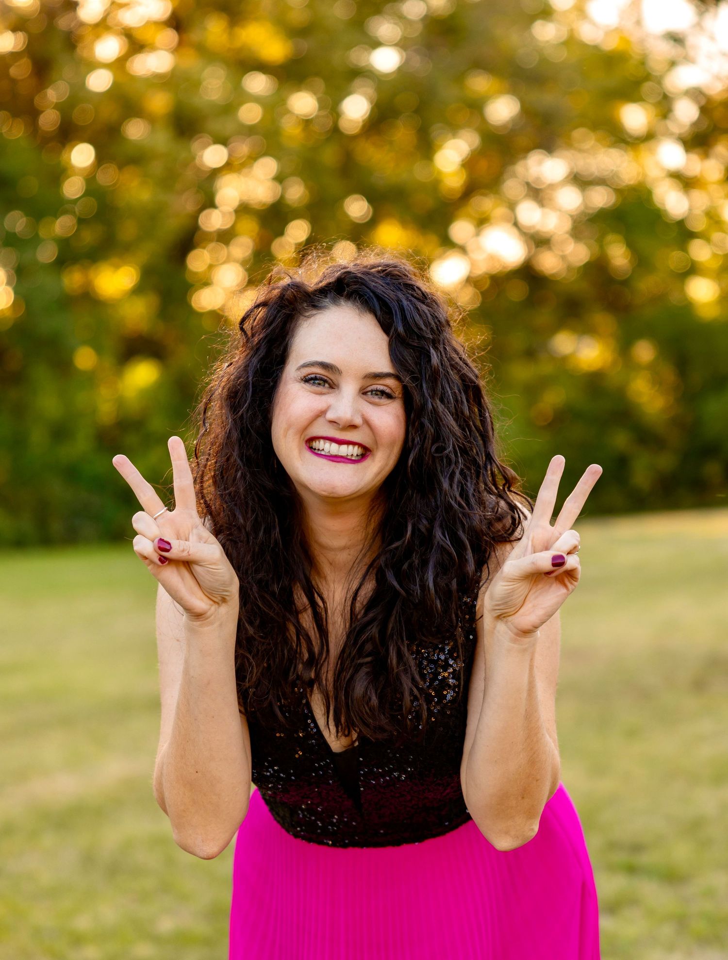A figure in a pink skirt and dark top poses outdoors making peace signs with both hands at sunset with bokeh background.