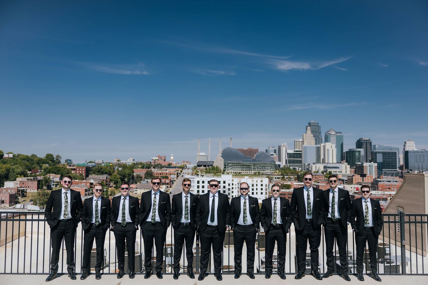 Group of groomsmen in matching black suits and ties stand in a line on rooftop with city skyline backdrop.