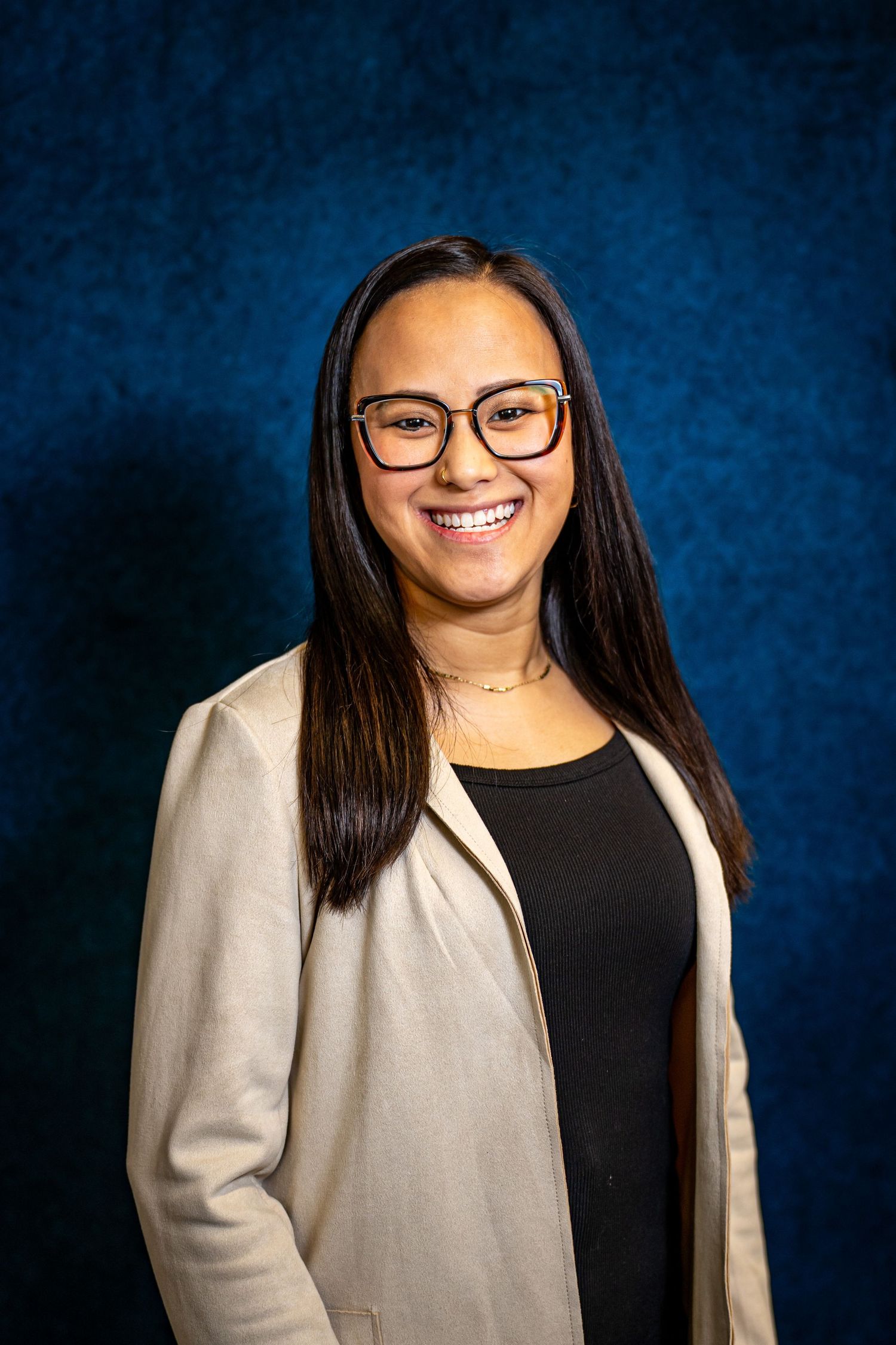 Smiling young woman wearing glasses and a beige blazer poses against a blue studio backdrop.