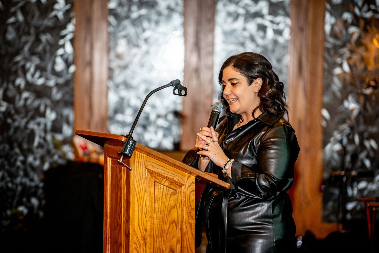Woman in black leather jacket speaking at wooden podium with microphone at formal event.