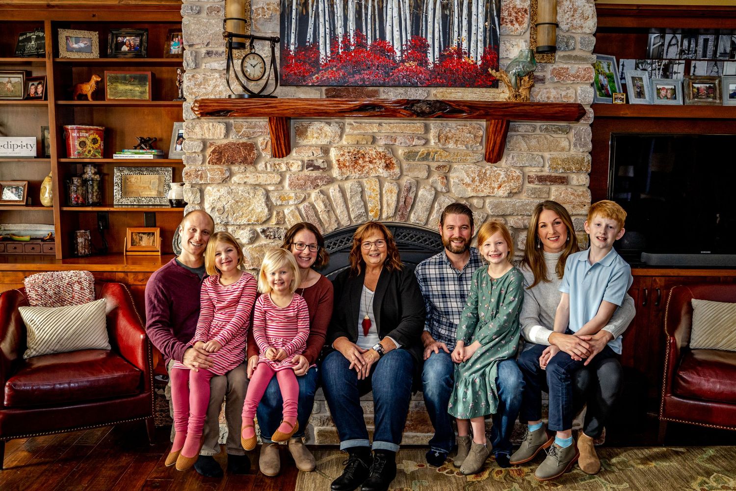 Large family group smiling together in a cozy living room with stone fireplace and wooden bookshelves.