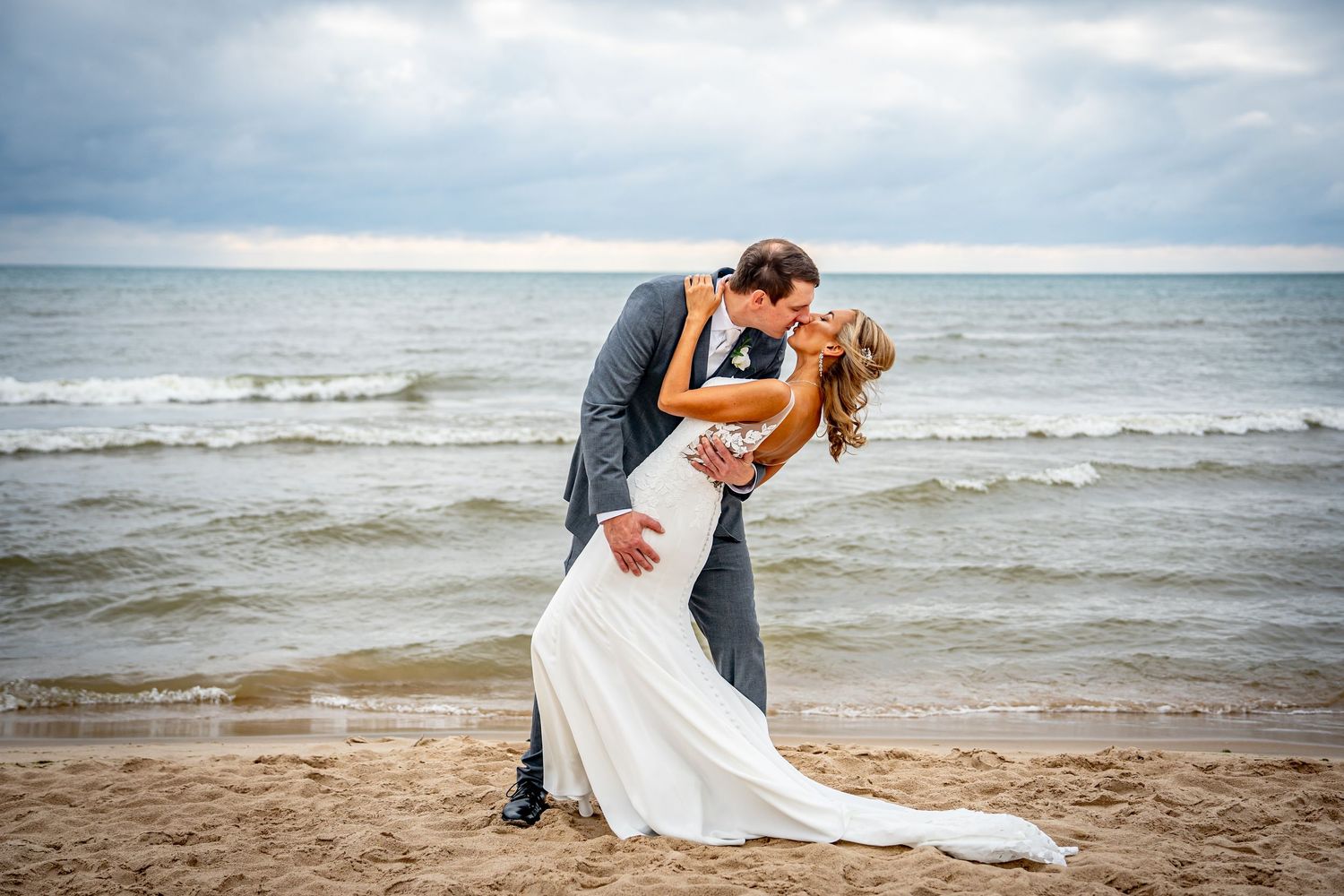 Bride and groom sharing a romantic dip kiss on a sandy beach with overcast skies behind them.