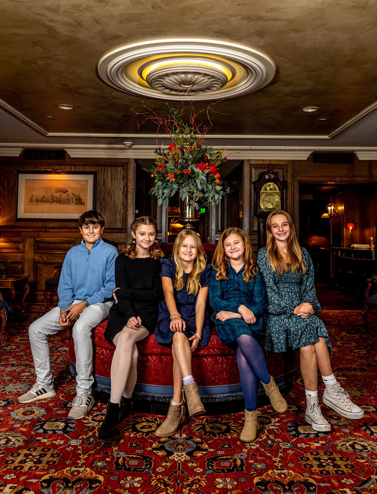 Five children sitting together on a red sofa in a festive decorated lobby with ornate ceiling.