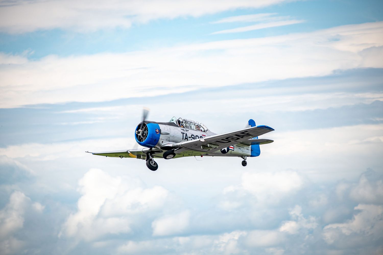 A vintage silver military aircraft with blue accents flies through a cloud-dotted sky during daytime.