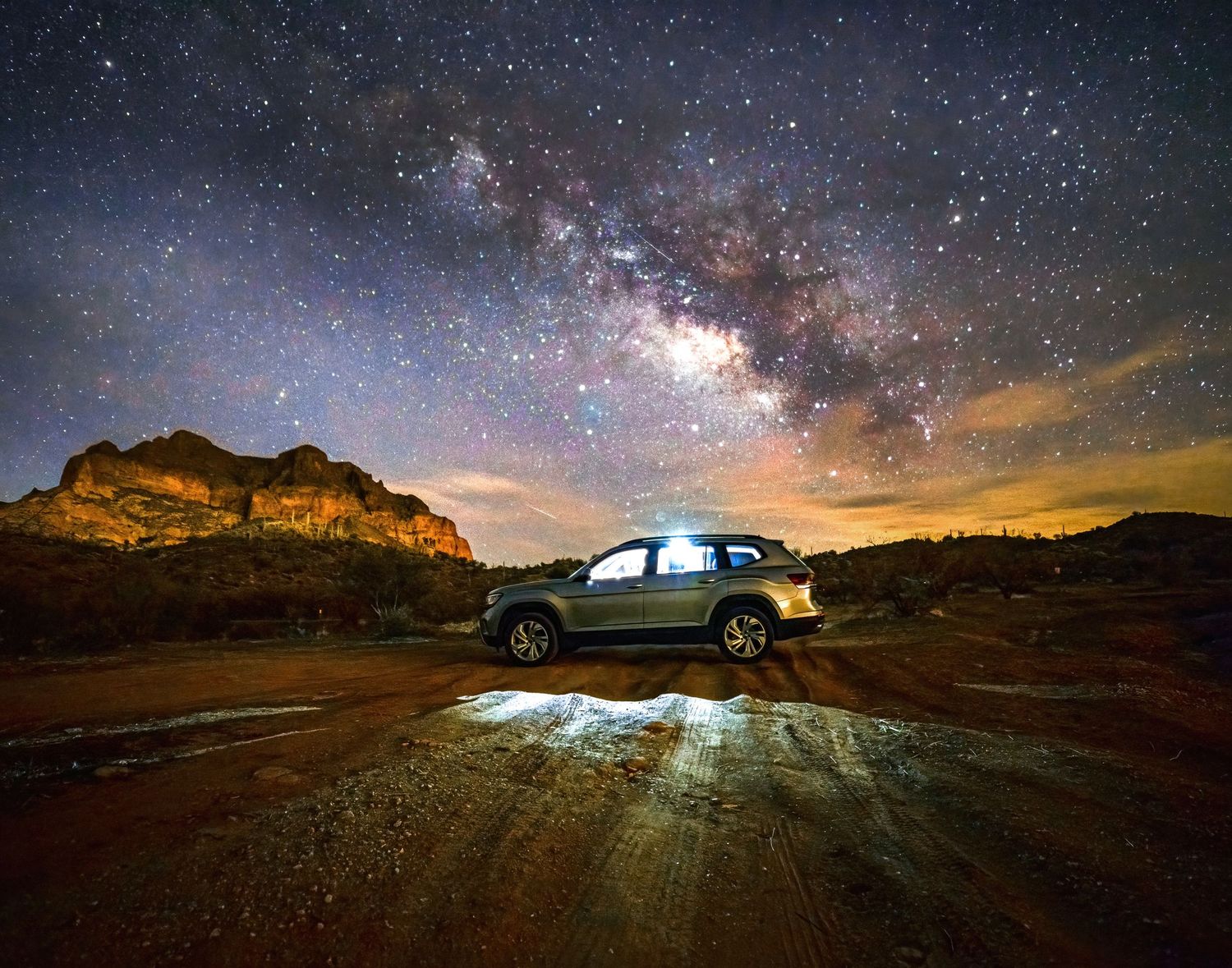 Silver SUV parked under brilliant Milky Way galaxy sky beside red rock formations at night.
