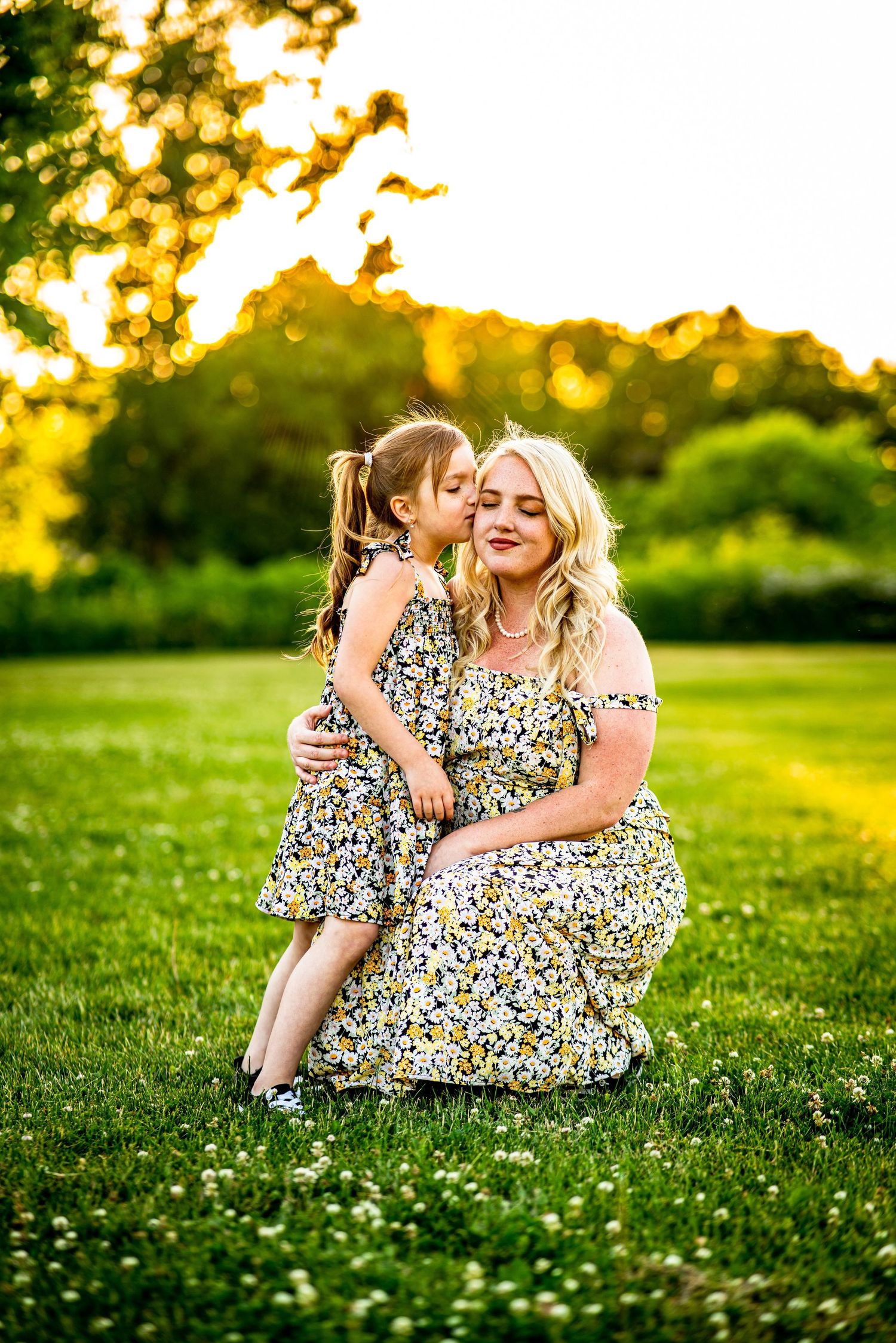 A mother and daughter in matching floral dresses sharing a tender moment in a sunlit green field.
