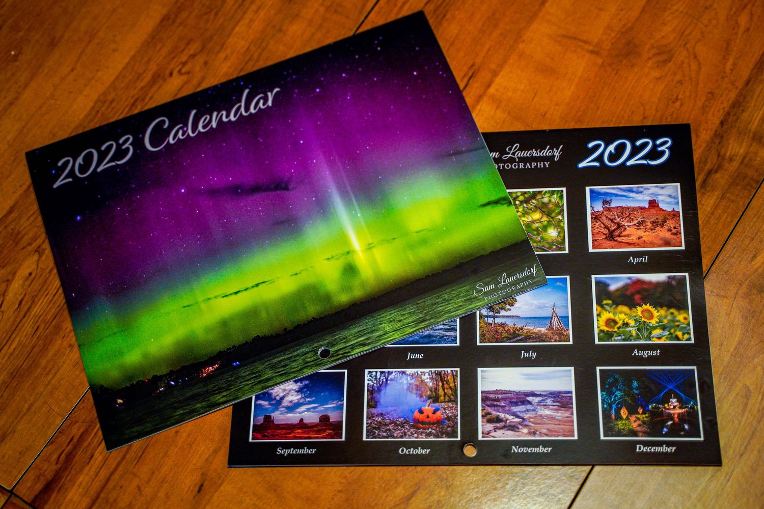 Two Sam Lauersdorf Photography 2023 calendars featuring aurora borealis cover on wooden surface.
