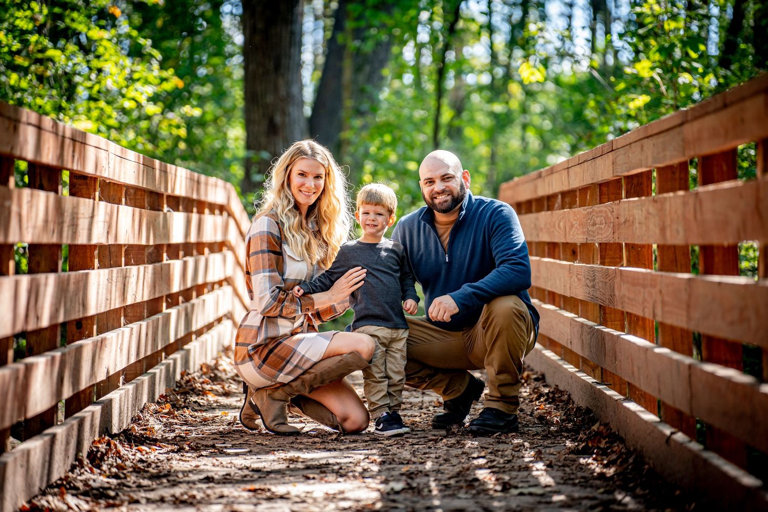 Happy family of three posing on a wooden bridge surrounded by autumn forest trees.