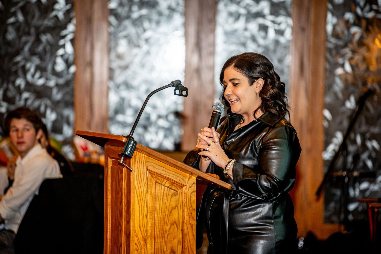 Woman in black leather jacket speaking at podium with microphone at indoor evening event.