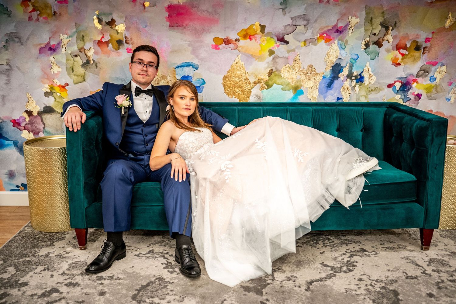 Bride and groom posing on teal velvet sofa in front of colorful abstract art backdrop at elegant wedding venue.