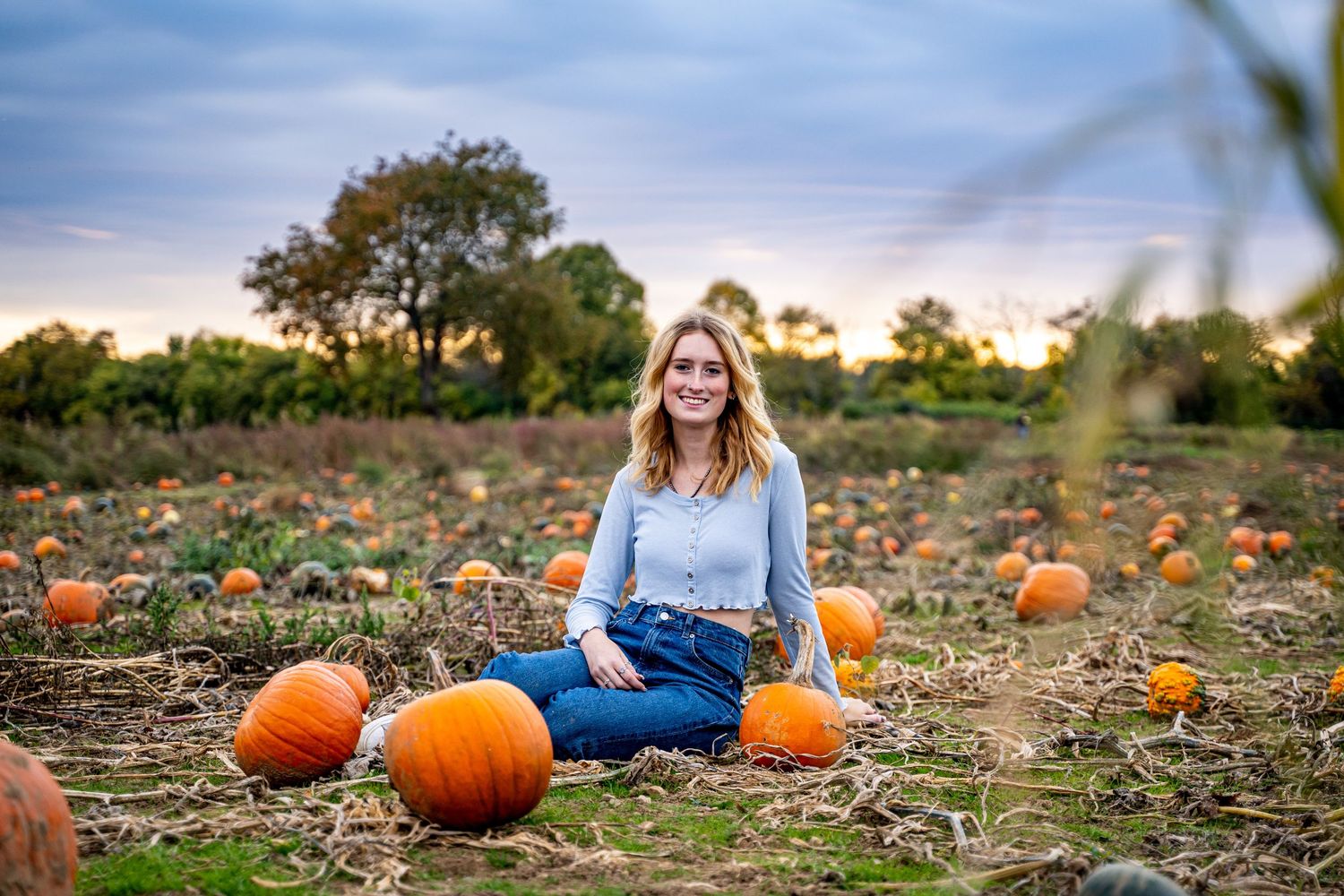 Young woman smiling in a pumpkin patch field at sunset, surrounded by orange pumpkins on a fall day.