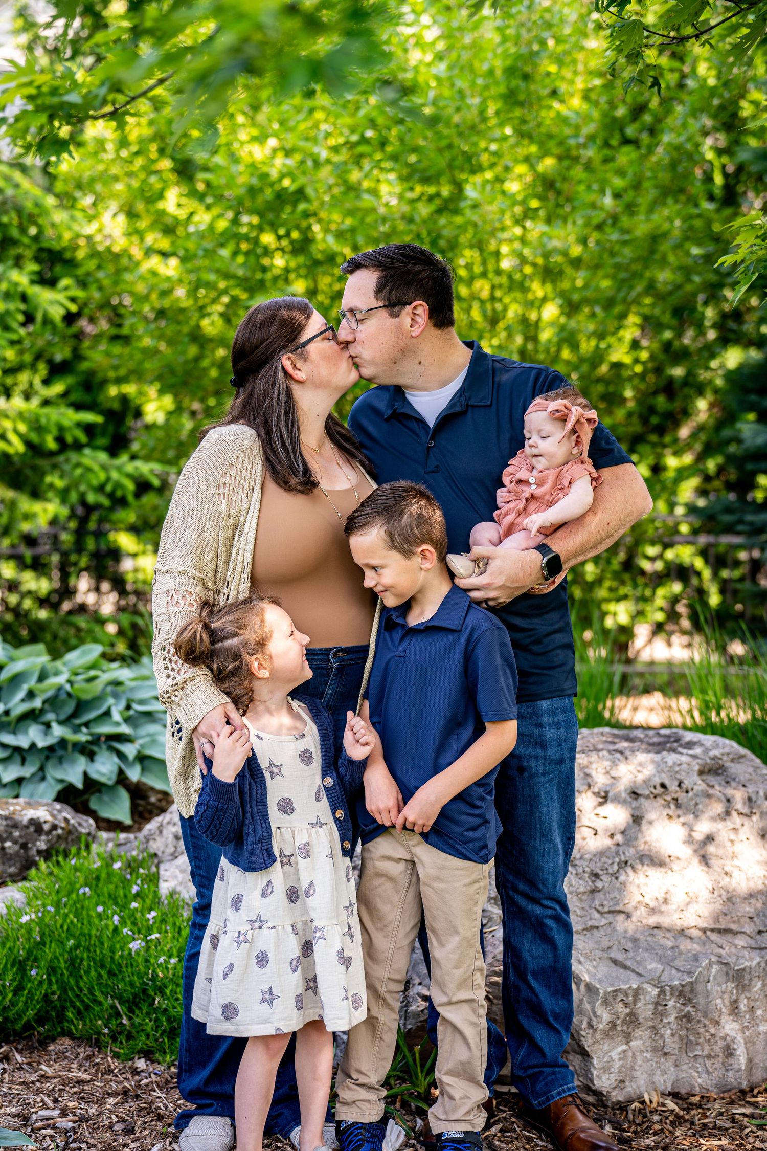 Family of five with kissing parents, two young children, and a baby in a lush green garden setting.