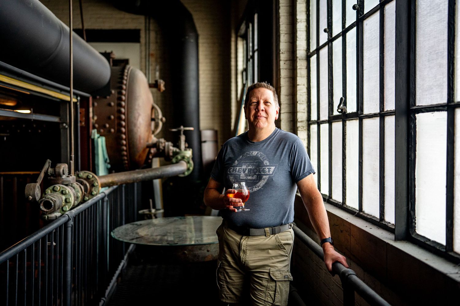 Person holding a craft beer glass in an industrial brewery setting with large machinery and warehouse windows.