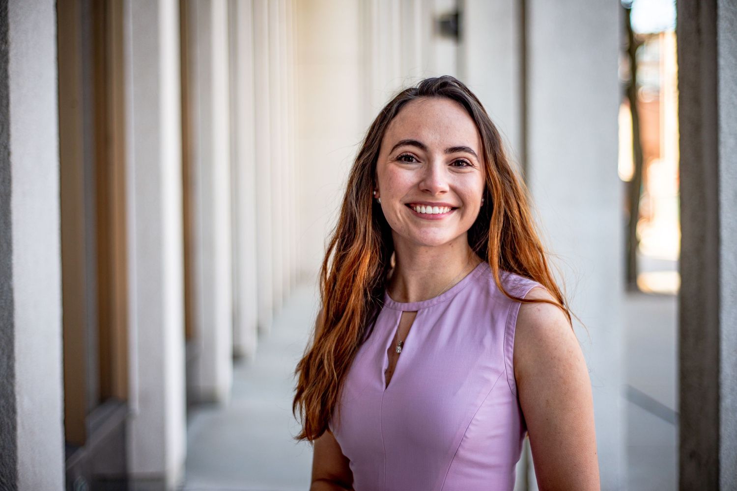 Smiling young woman with long auburn hair wearing a lavender sleeveless top, standing between white columns.