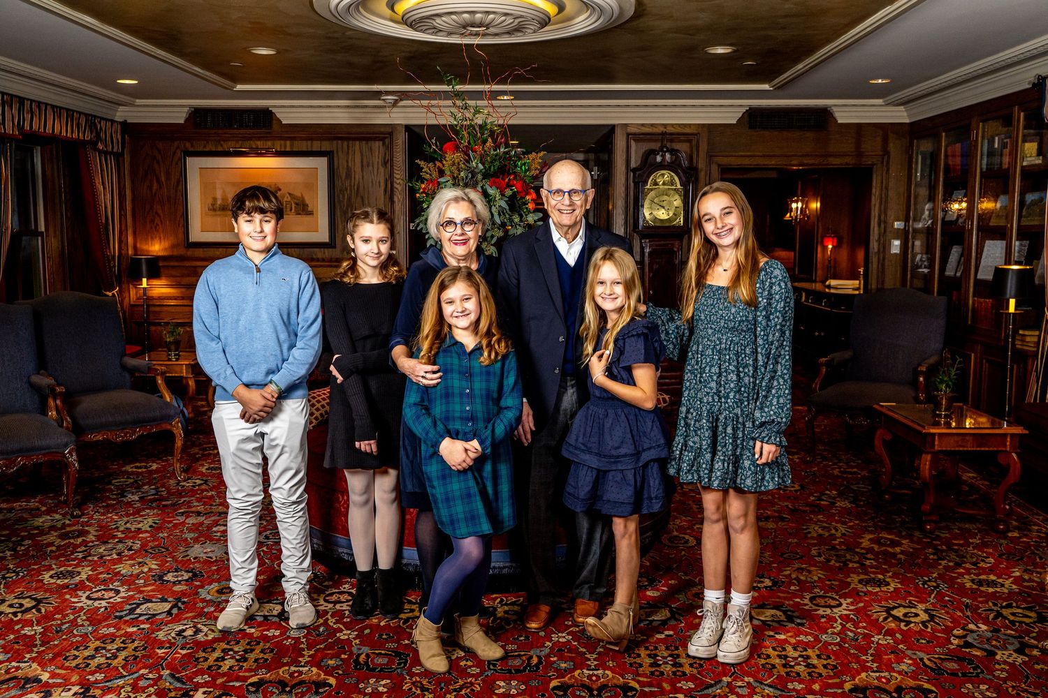 Family of seven poses together in an elegant room decorated for Christmas, with a grandfather clock and holiday tree in background.