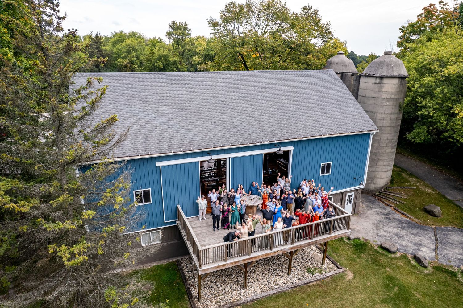 Aerial view of a blue barn venue with guests on a deck, surrounded by trees and a silo.