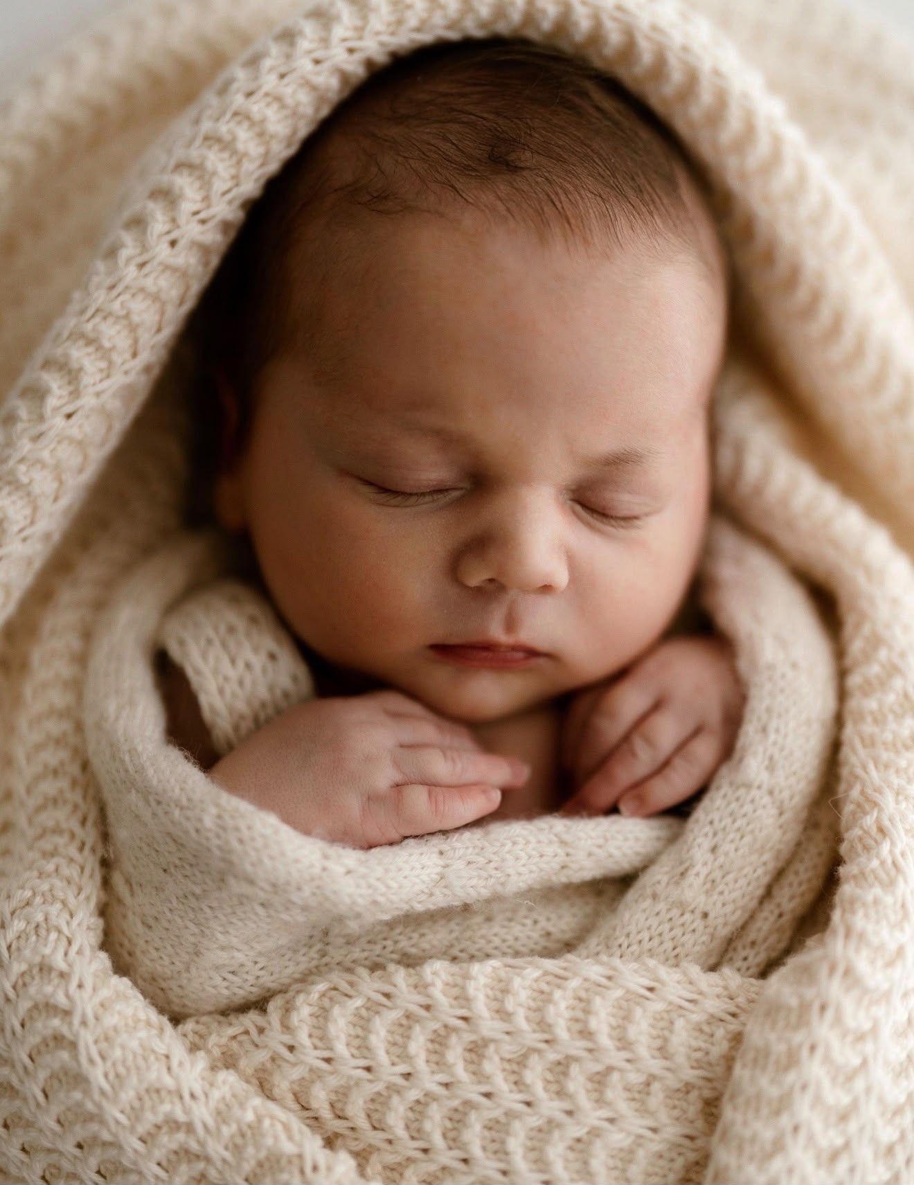 A peaceful sleeping newborn wrapped in a cream blanket, with tiny hands resting gently against their chest in a timeless, neutral-tones.
