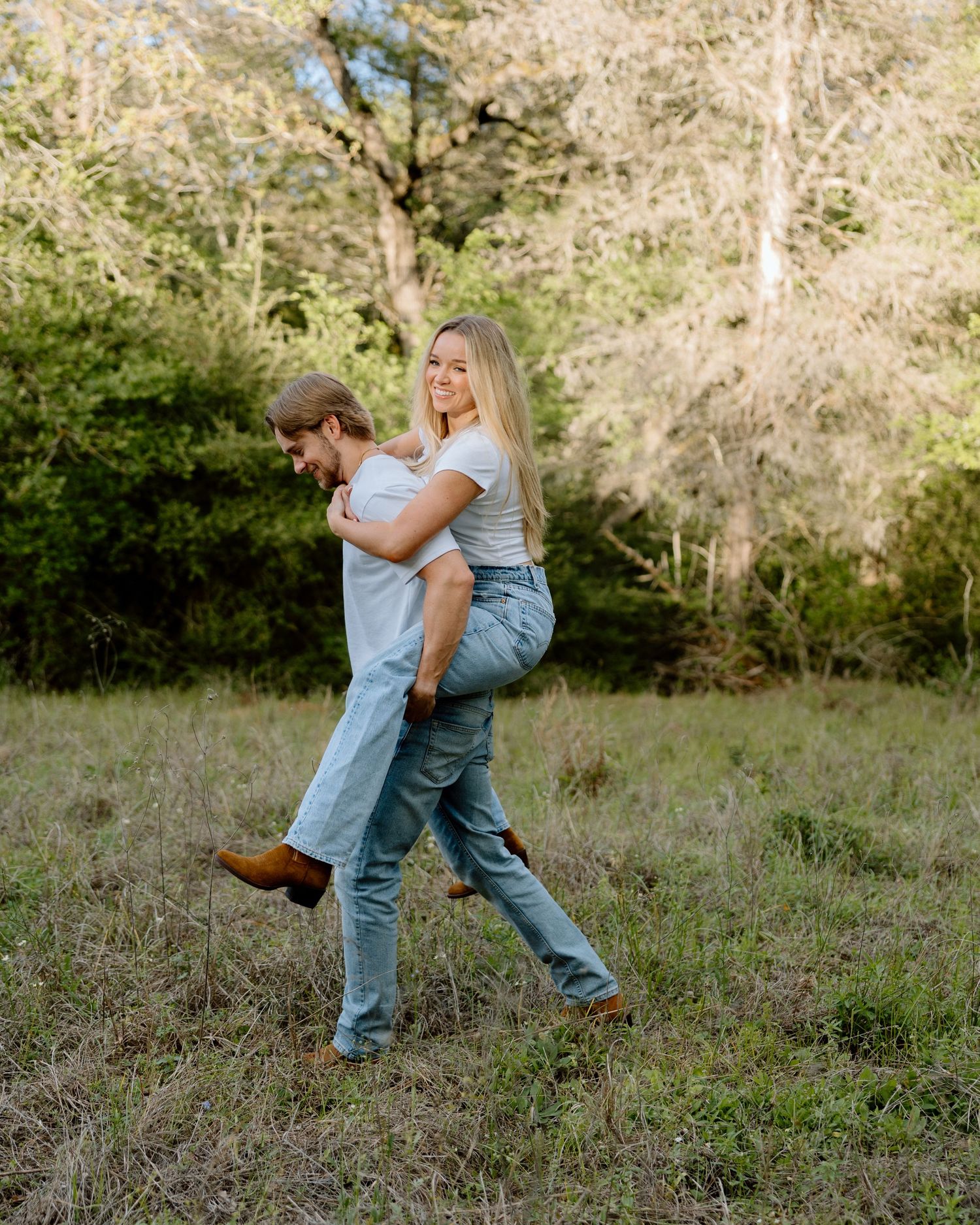 A man carries a smiling blonde woman on his back in a grassy field surrounded by lush green trees.
