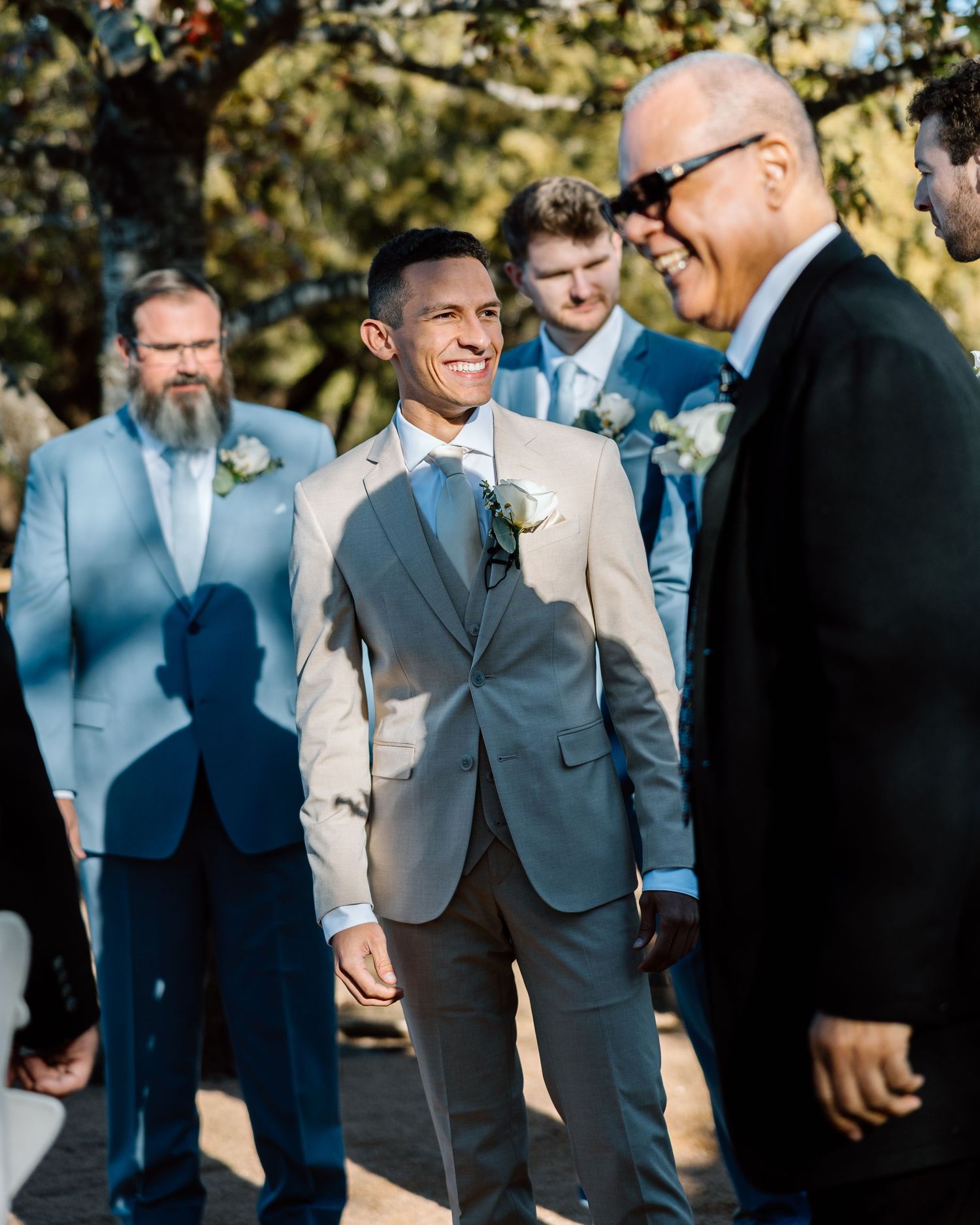 Smiling groom in beige suit standing with groomsmen in blue suits outdoors.